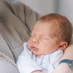 Paula Jankowska Photography - Śpiący noworodek w białym ubranku, trzymany w ramionach osoby w beżowej koszuli. Delikatne światło podkreśla rysy twarzy dziecka.