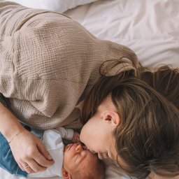 Paula Jankowska Photography - Matka w beżowej bluzce całuje śpiącego noworodka w białym ubranku, leżąc na białej pościeli. Delikatne światło podkreśla intymność i bliskość relacji.