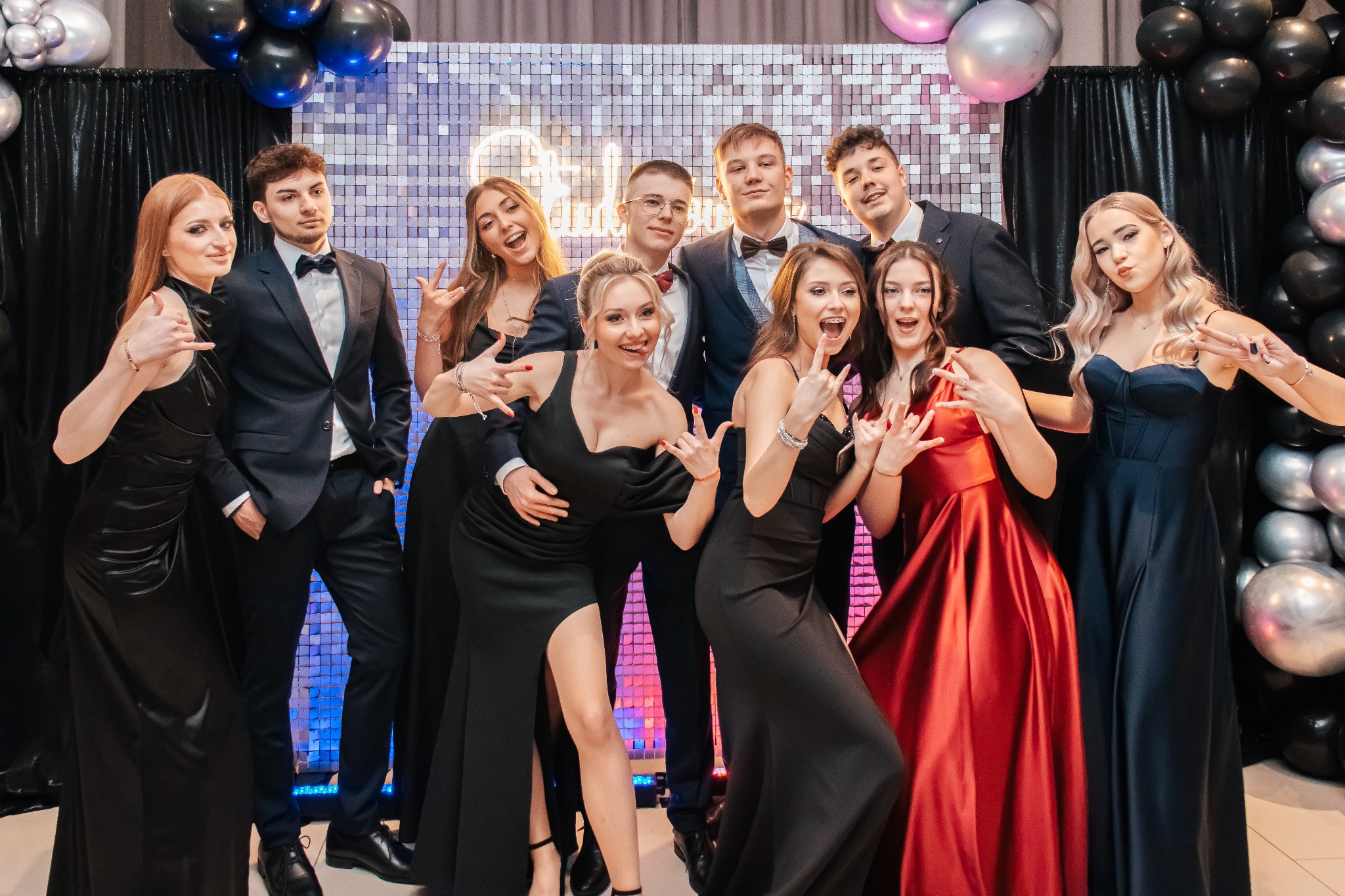 Grupa młodych, elegancko ubranych osób pozuje na tle błyszczącej ściany z balonami, wykonując gest dłońmi.