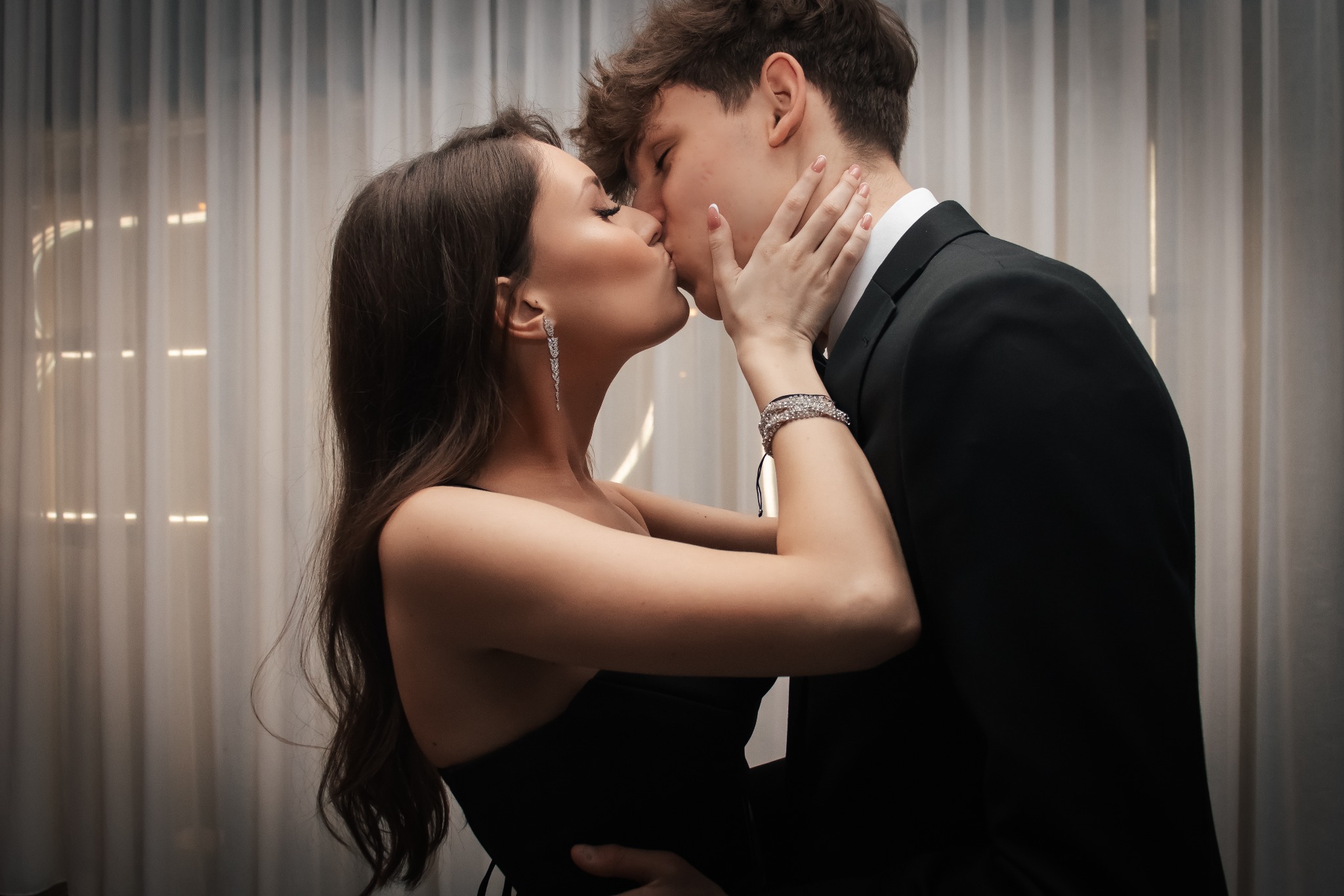 Elegancka para całująca się na tle delikatnych, białych zasłon; kobieta w czarnej sukience z diamentowymi kolczykami i bransoletką, mężczyzna w czarnym garniturze.
