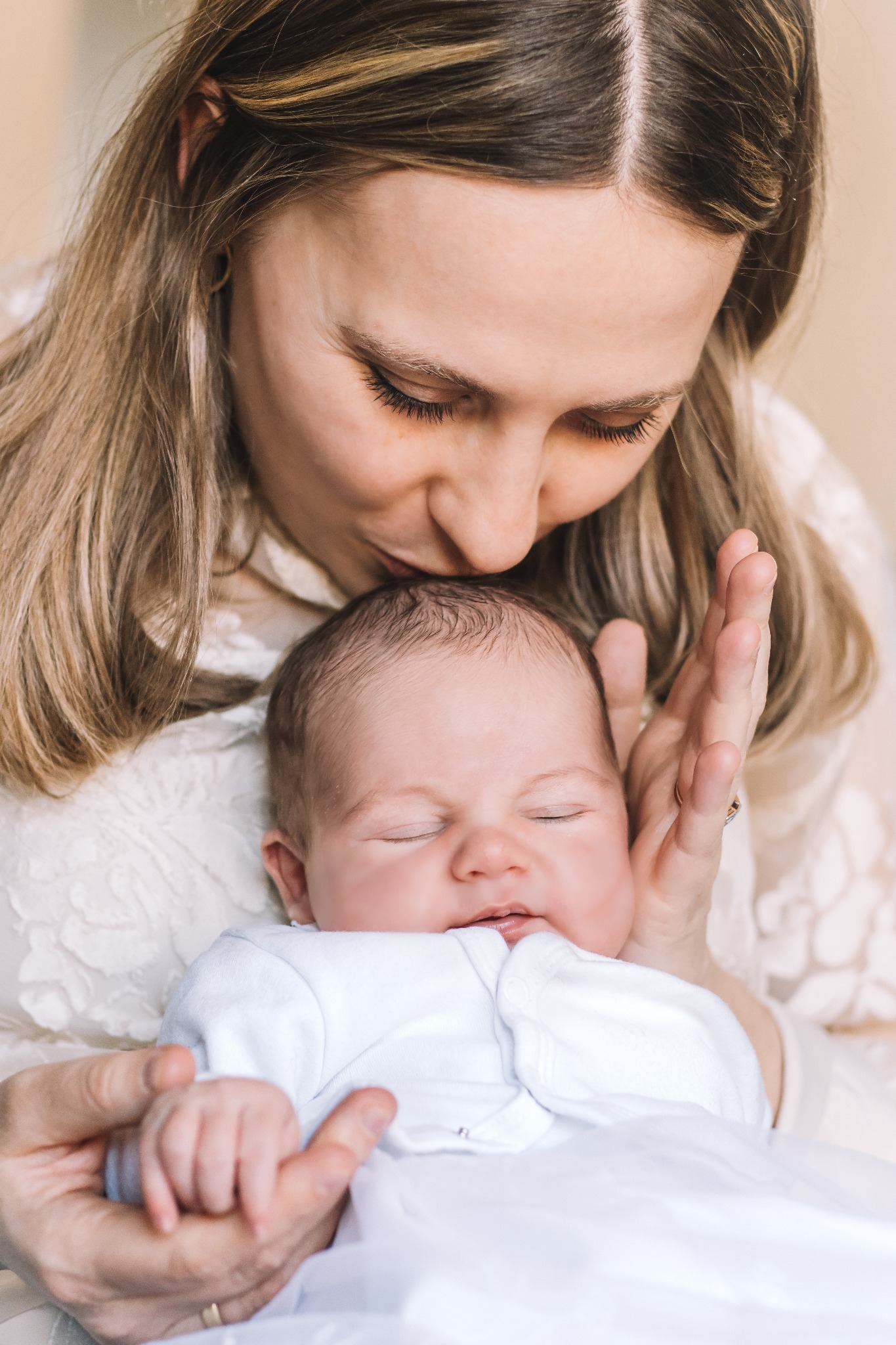 Matka całuje śpiącego noworodka, delikatnie trzymając go w dłoniach, ubrana w koronkową bluzkę.