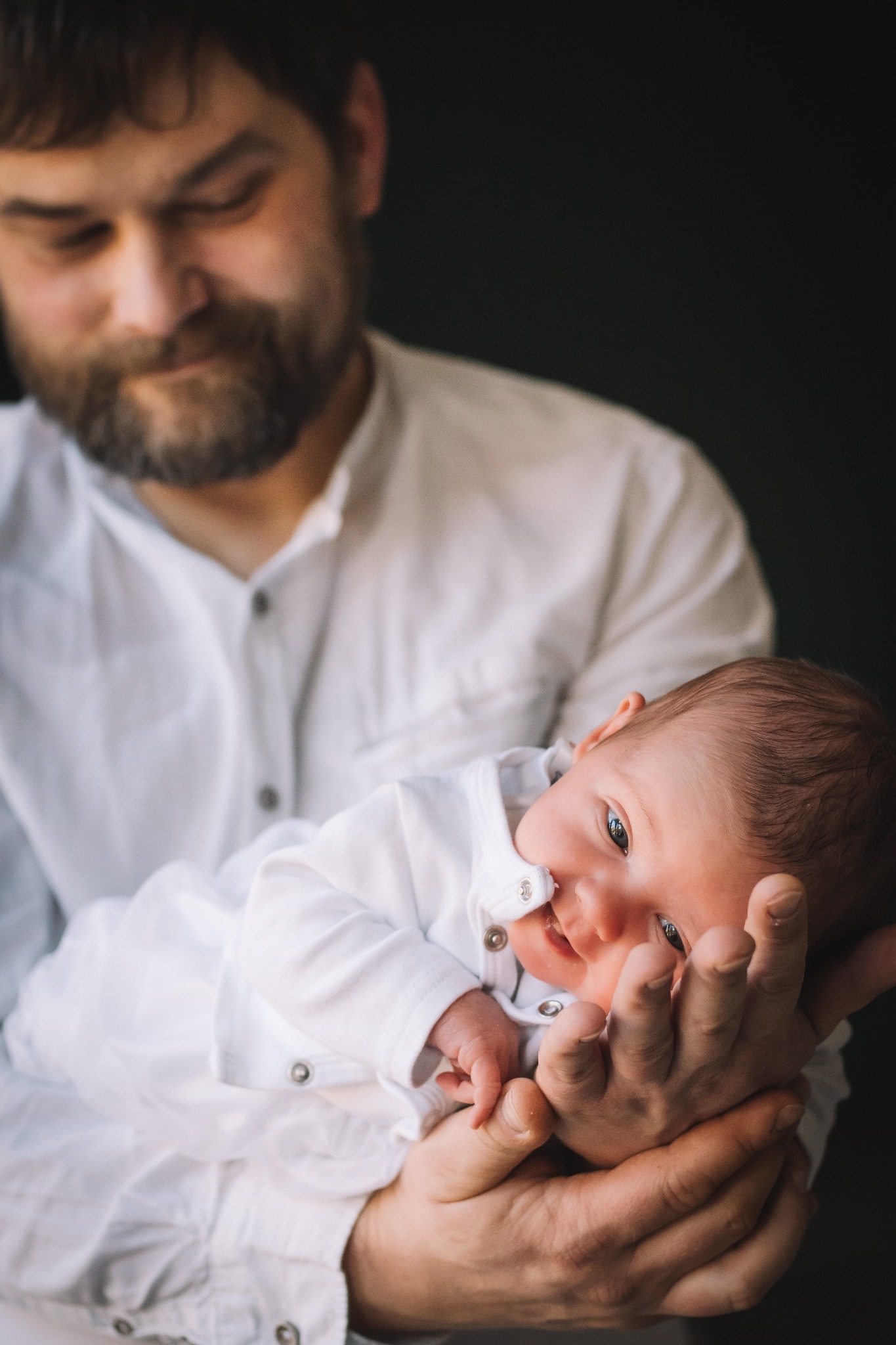 Noworodek w białym ubranku, trzymany w dłoniach przez brodatego mężczyznę w białej koszuli, ciemne tło.