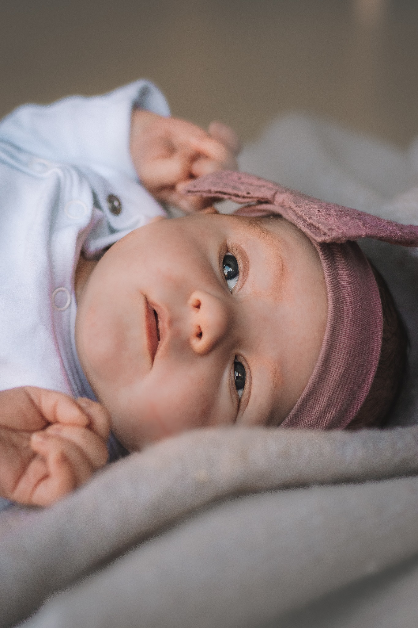 Noworodek leżący na miękkim kocyku, ubrany w białe body i różową opaskę z koronką, patrzy w górę z delikatnym uśmiechem.