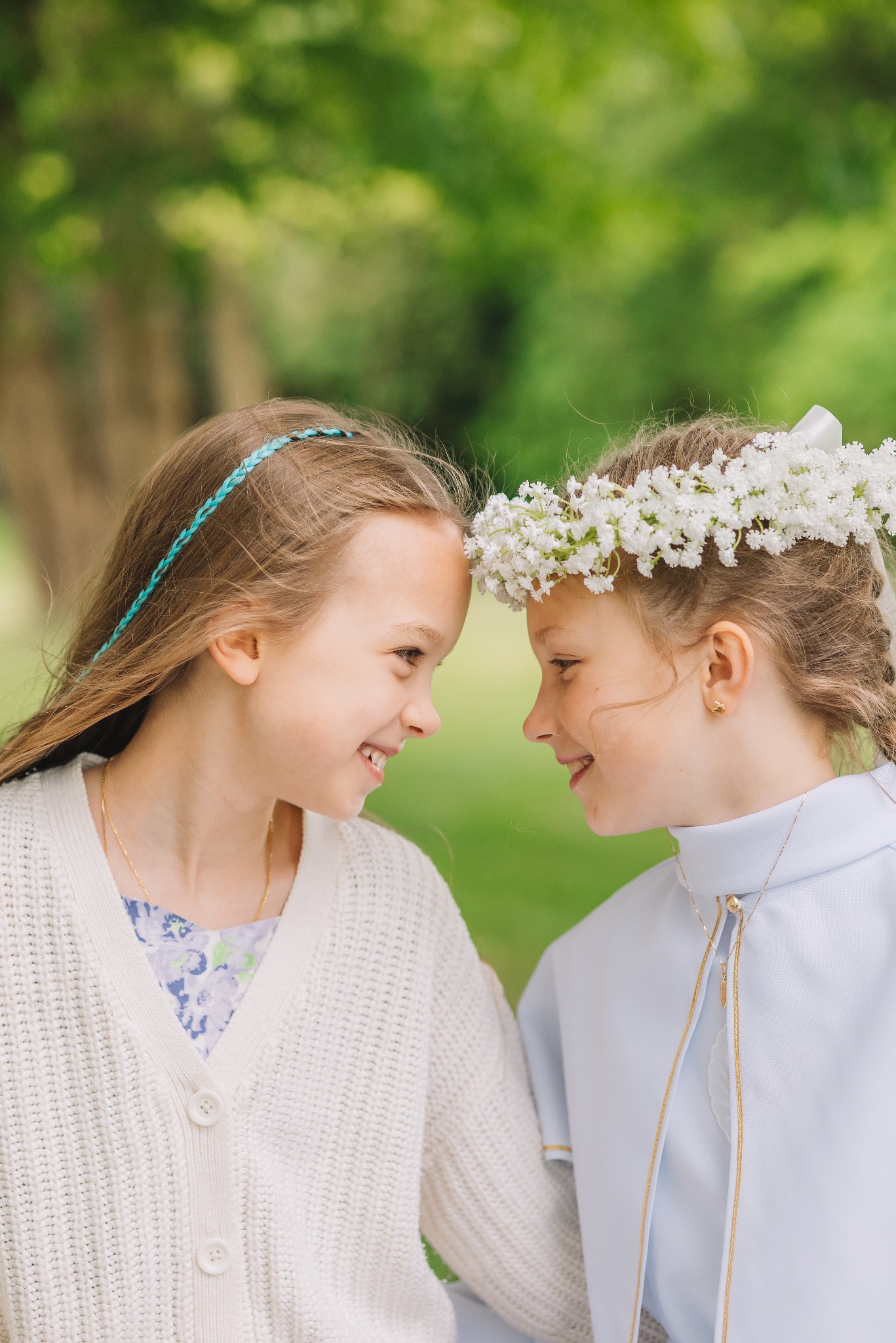 Portret dwóch uśmiechniętych dziewczynek, jedna w wianku z białych kwiatów i albie komunijnej, druga w swetrze, stojących blisko siebie na tle zieleni.