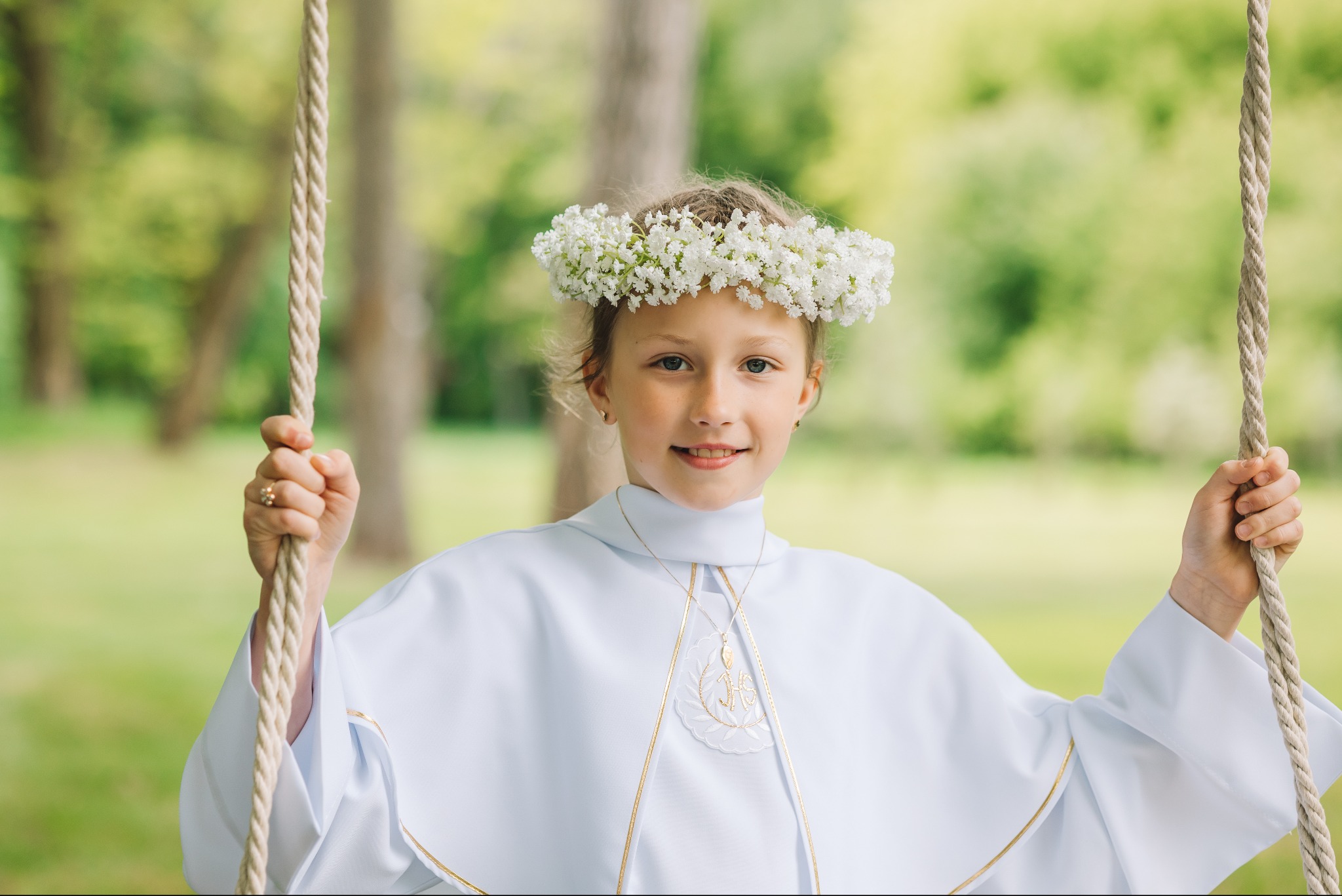 Dziewczynka w białej albie i wianku z białych kwiatów na huśtawce linowej, uśmiechnięta, z delikatnym makijażem, na tle zieleni drzew.