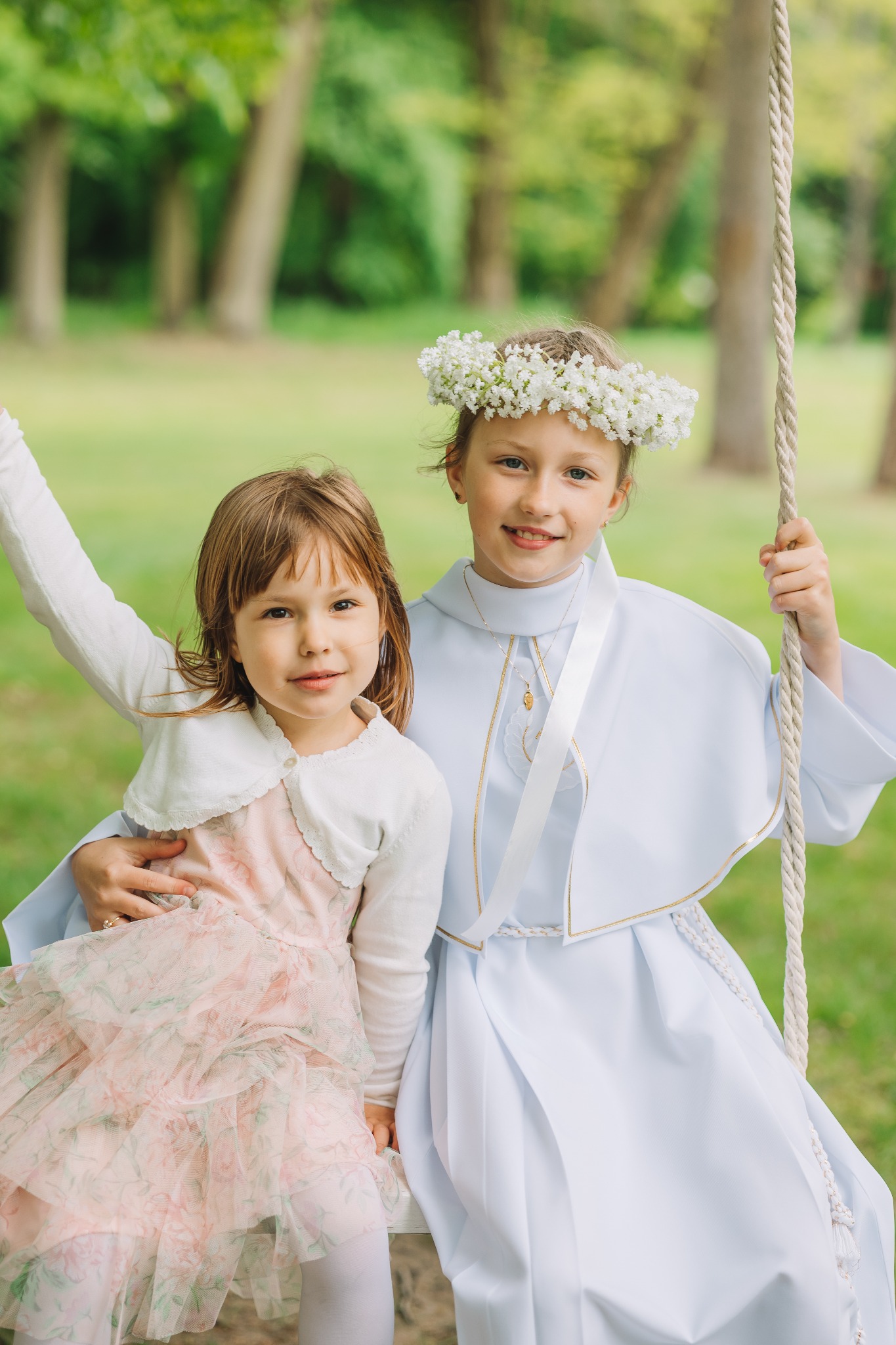 Dwie dziewczynki na huśtawce w plenerze. Jedna w stroju komunijnym z wiankiem, druga w sukience w kwiaty. Uśmiechnięte, w naturalnym oświetleniu.