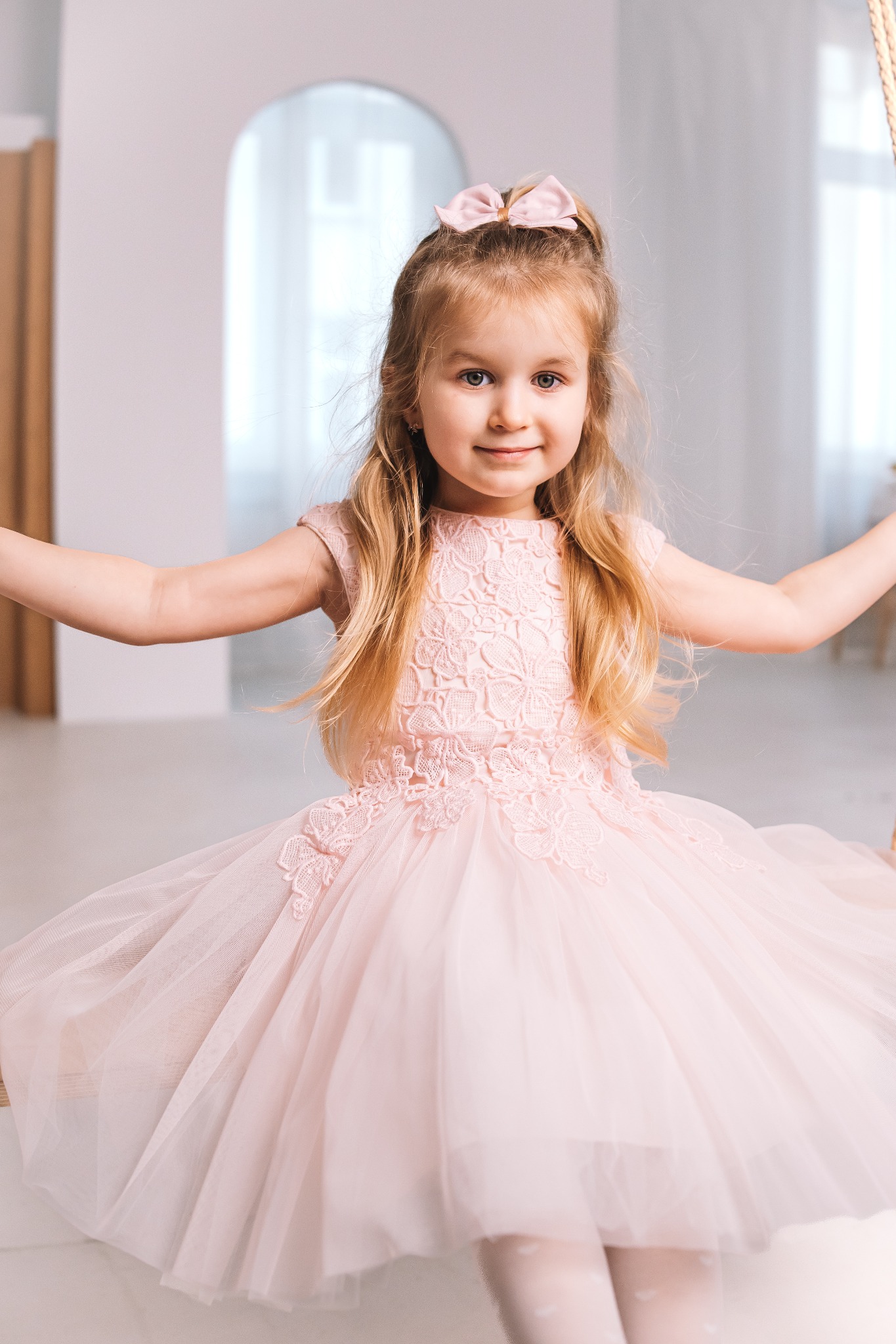 Urocza dziewczynka w pudrowej sukience z kokardą we włosach, siedząca na huśtawce w jasnym wnętrzu. Delikatne światło, pastelowa kolorystyka, subtelny uśmiech dziecka.