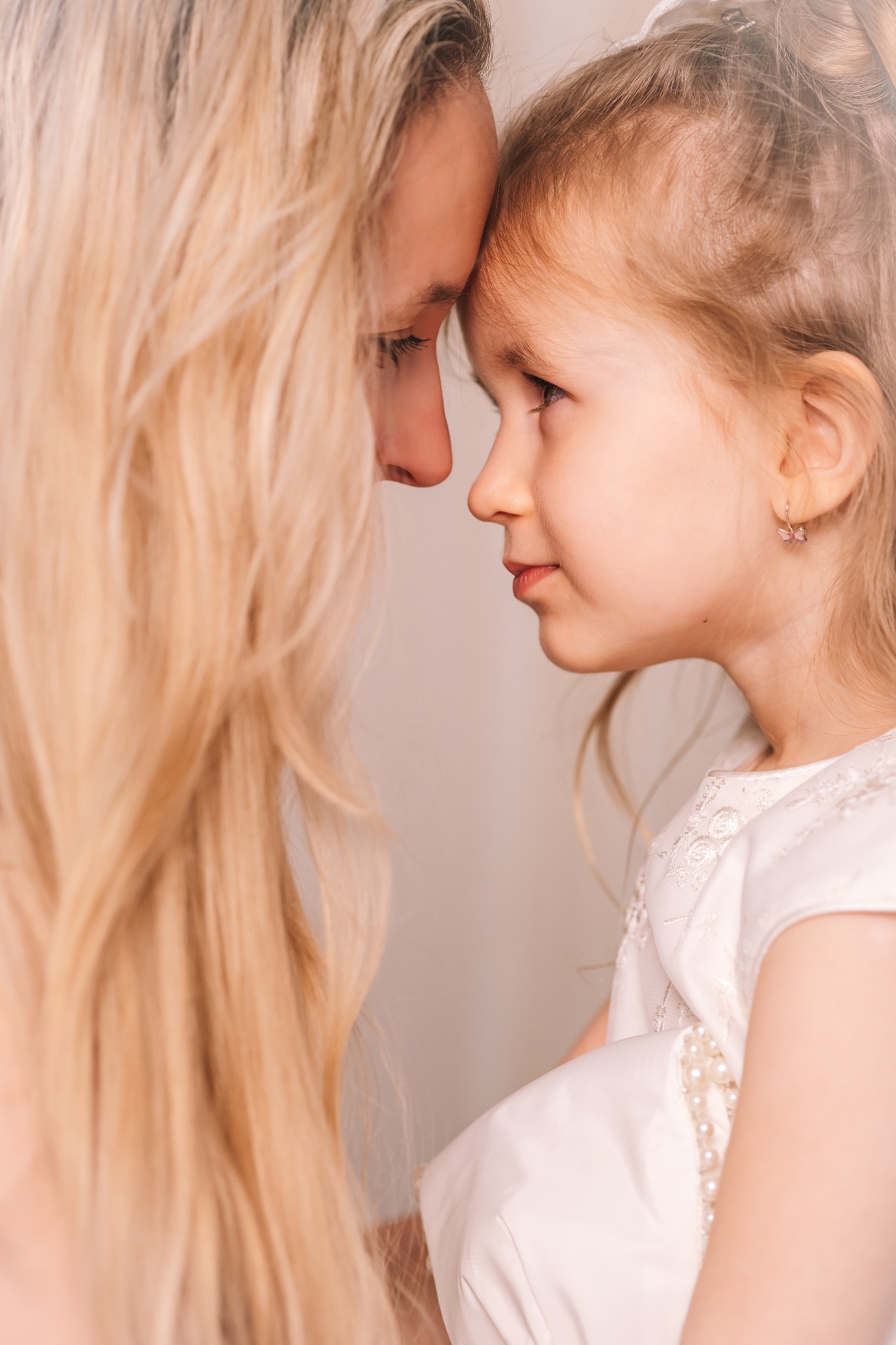 Profilowe zbliżenie matki z długimi blond włosami i córki z delikatnymi kolczykami, stykających się czołami, w miękkim, rozproszonym świetle.