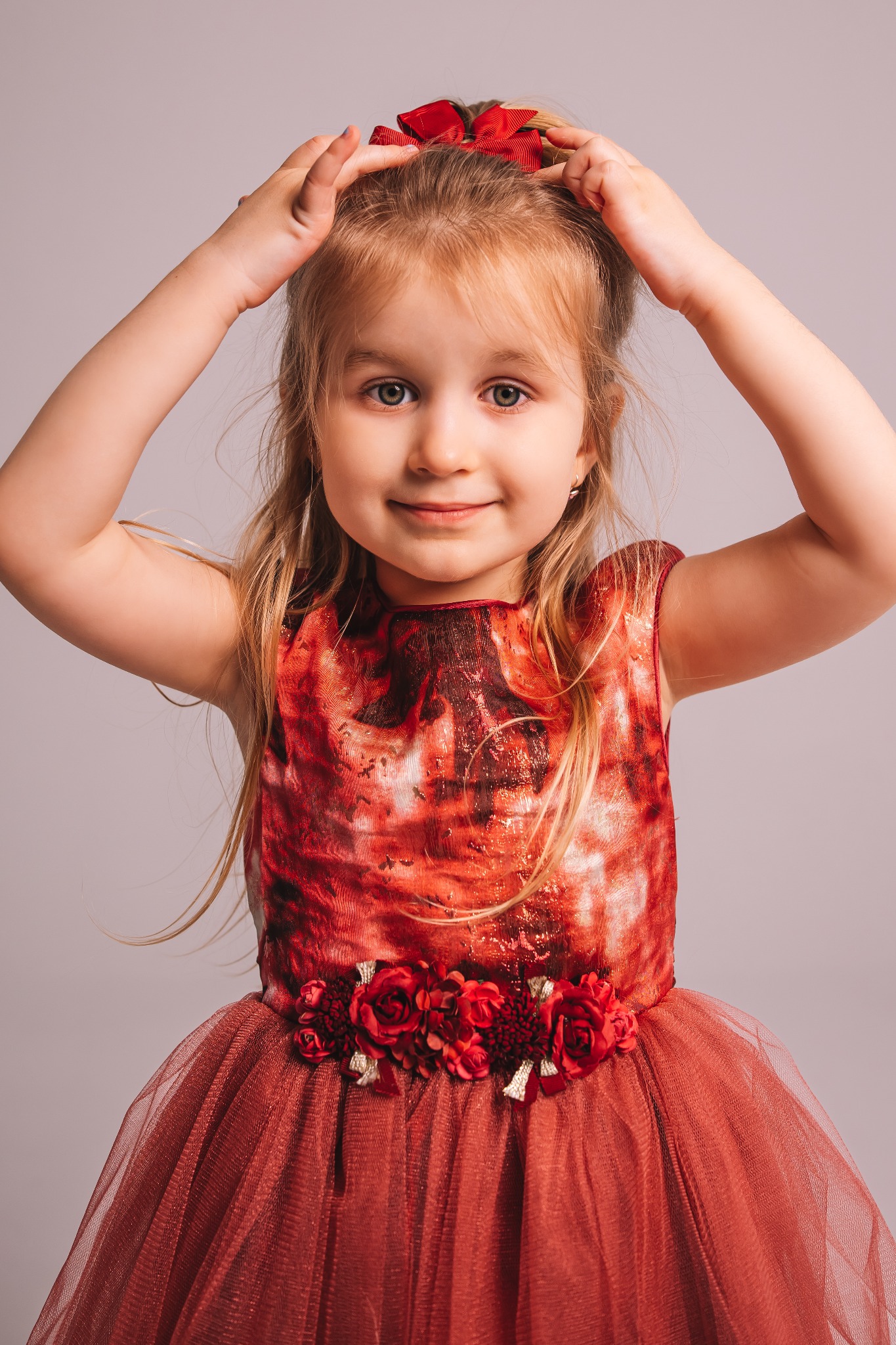 Portret uśmiechniętej dziewczynki w czerwonej sukience z tiulowym dołem i kwiatowym pasem, z czerwoną kokardą we włosach na jasnym tle.