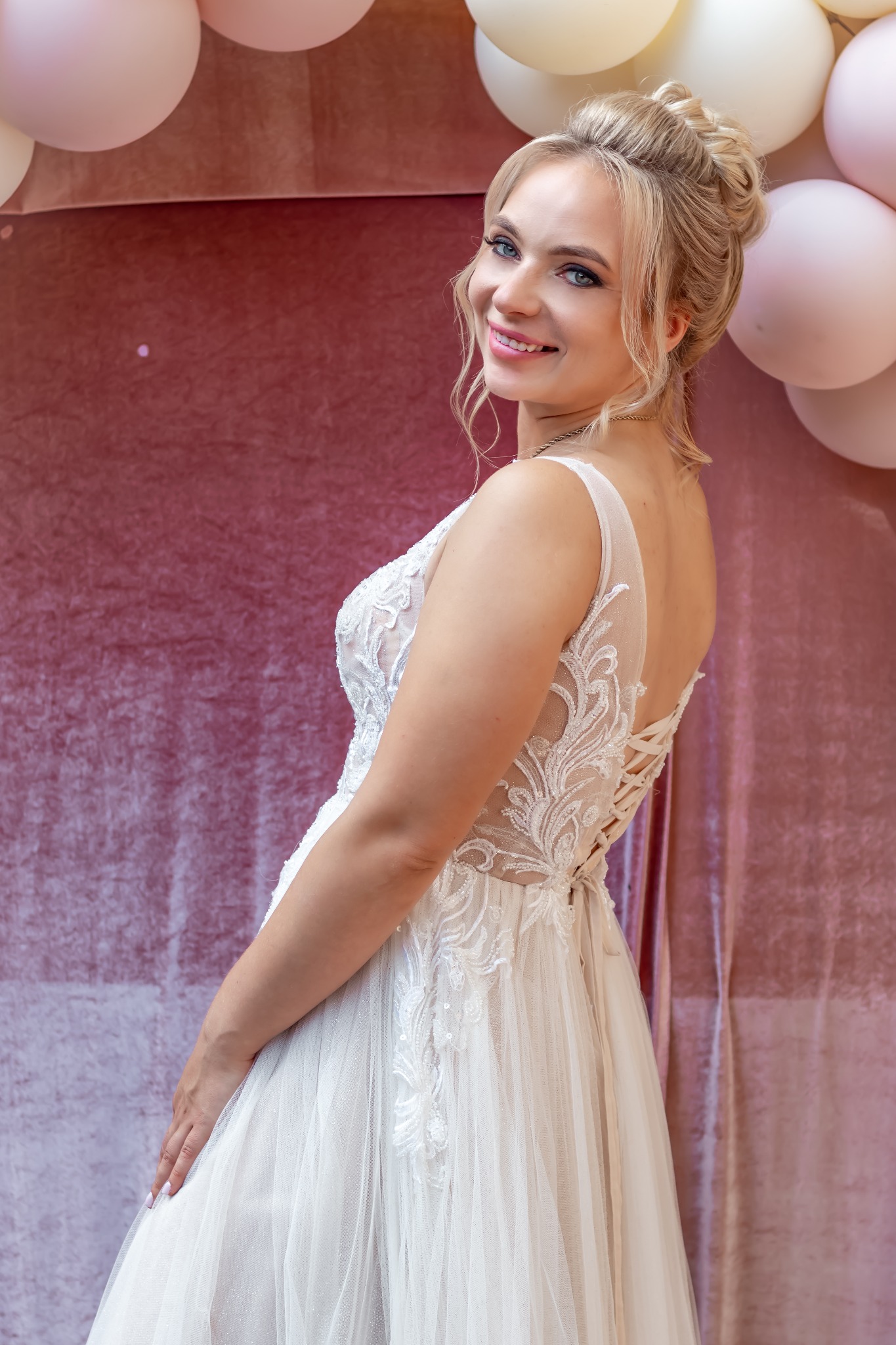 Uśmiechnięta panna młoda w koronkowej sukni, z upiętymi blond włosami, pozuje na tle różowej ściany z balonami. Widok zza ramienia, delikatny makijaż.