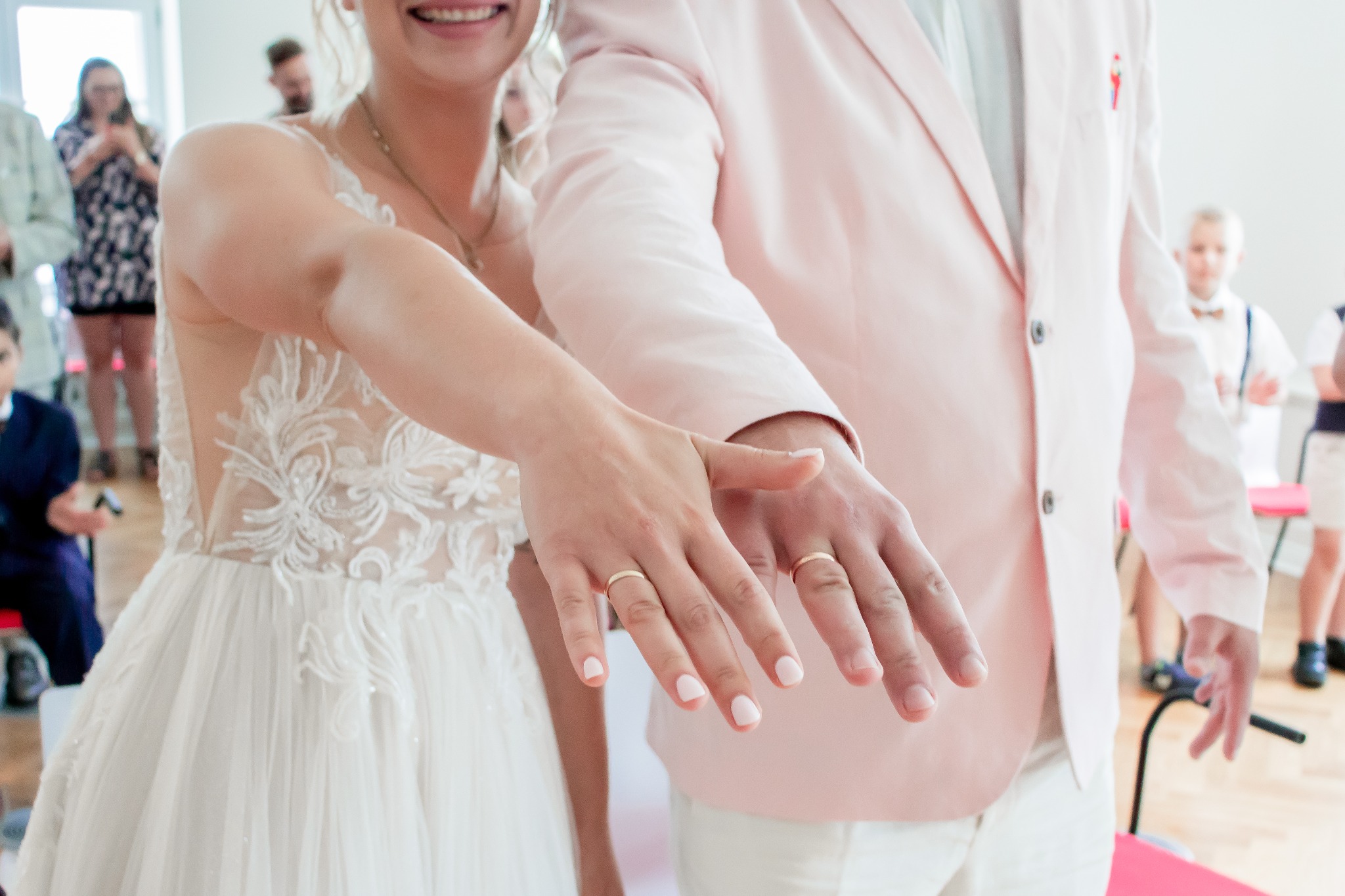 Nowożeńcy prezentują obrączki na palcach, ona w koronkowej sukni, on w różowym garniturze. W tle goście weselni, jasne wnętrze.