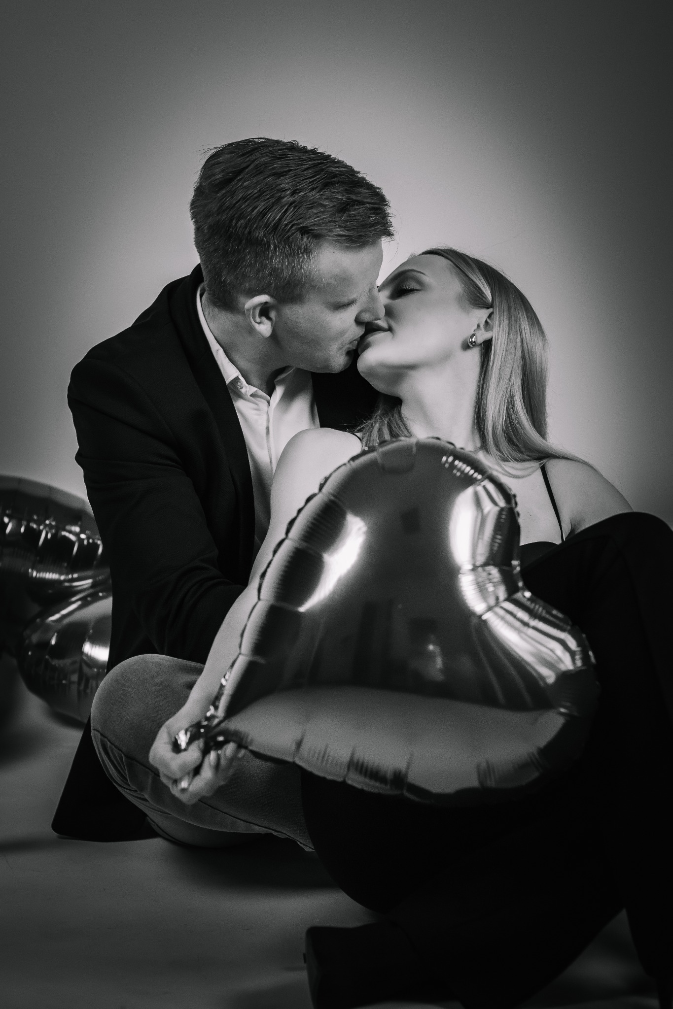 Czarno-białe zdjęcie całującej się pary z balonami w kształcie serc. Mężczyzna w marynarce, kobieta w sukience na tle szarego studia. Romantyczny, intymny moment.