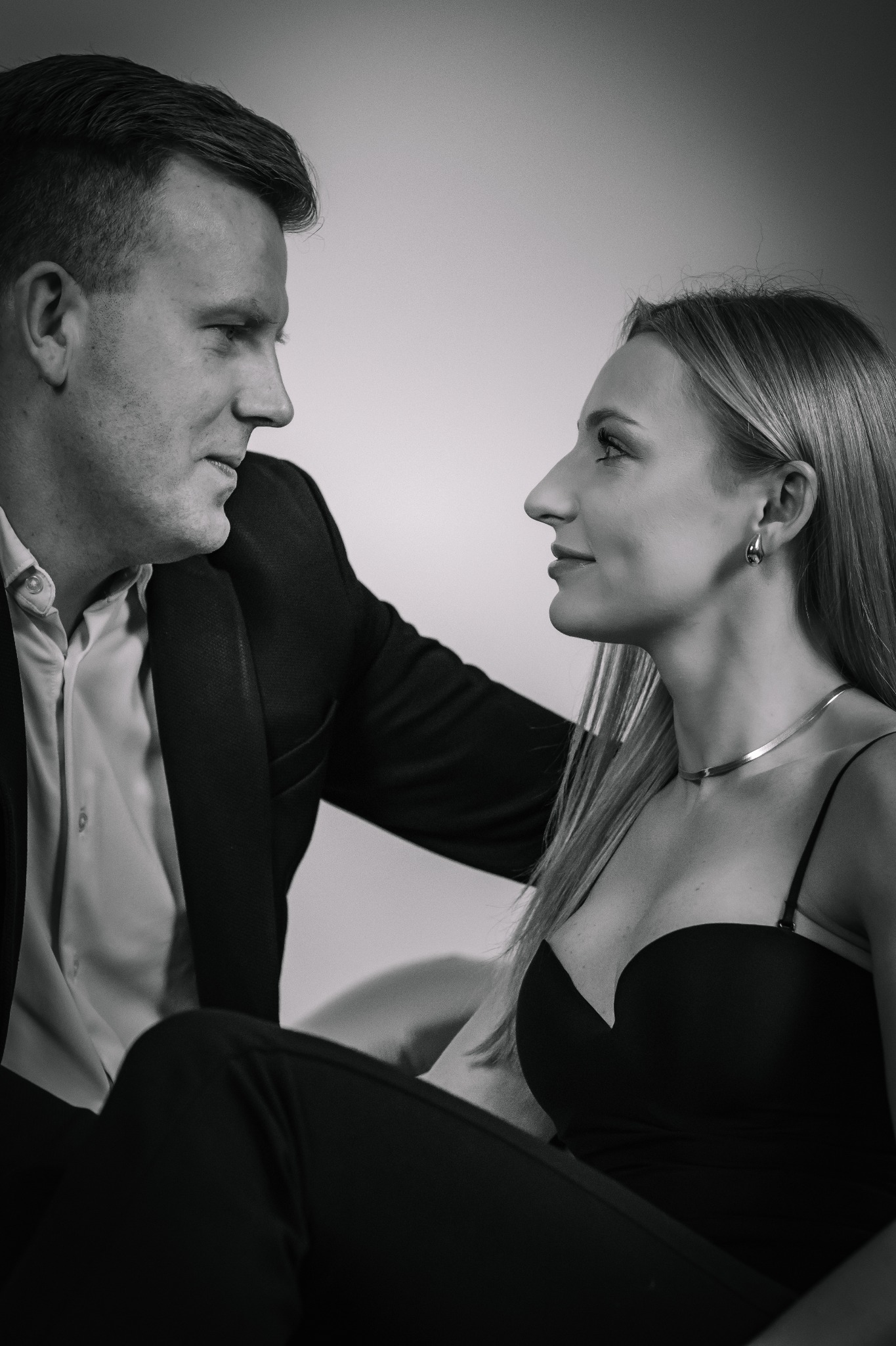 Czarno-białe, profilowe ujęcie eleganckiej pary. Mężczyzna w garniturze spogląda na kobietę w sukience z dekoltem. Delikatne oświetlenie, minimalistyczne tło.