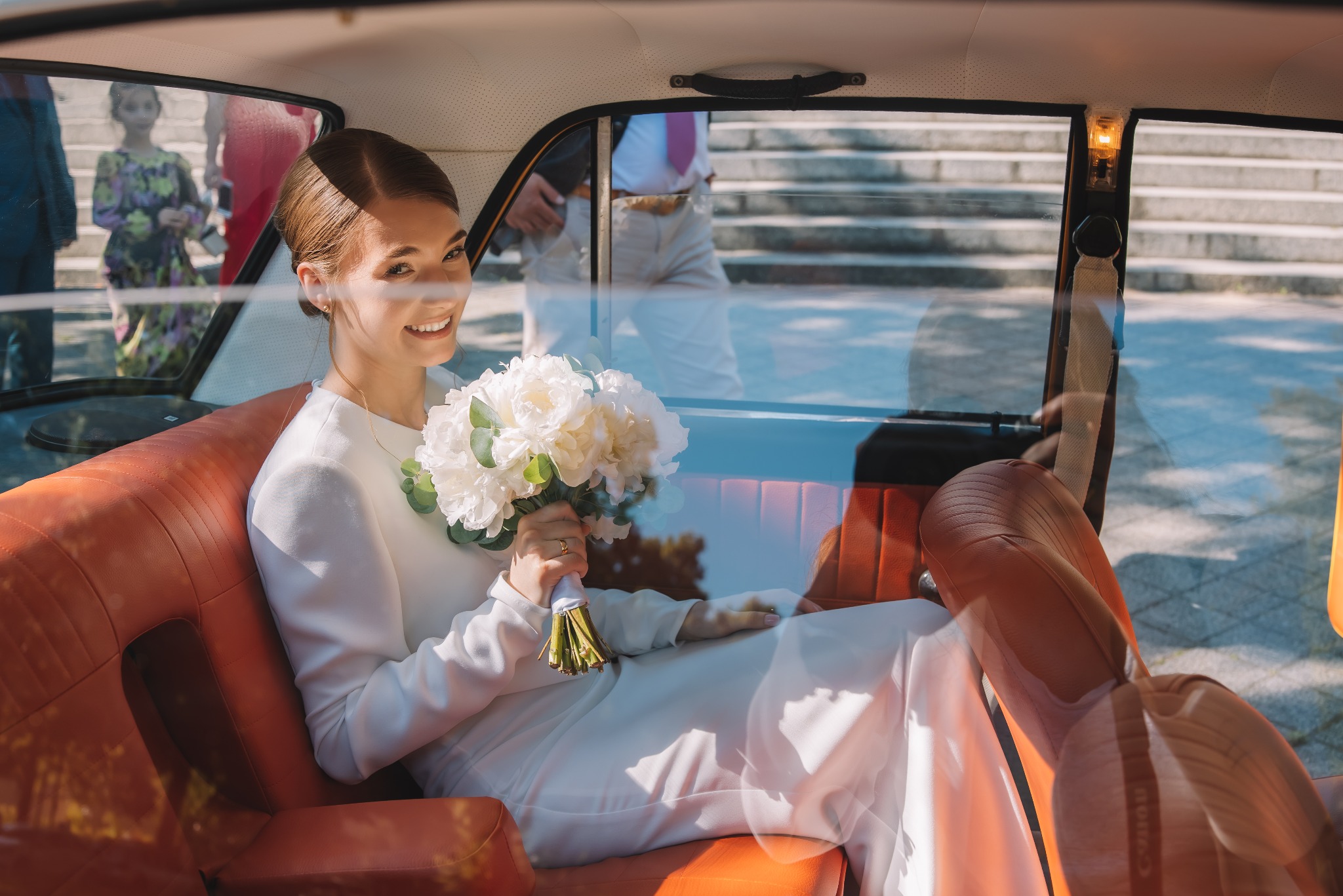 Uśmiechnięta panna młoda w białej sukni siedzi w samochodzie z bukietem białych kwiatów. W tle widoczni goście weselni i schody.