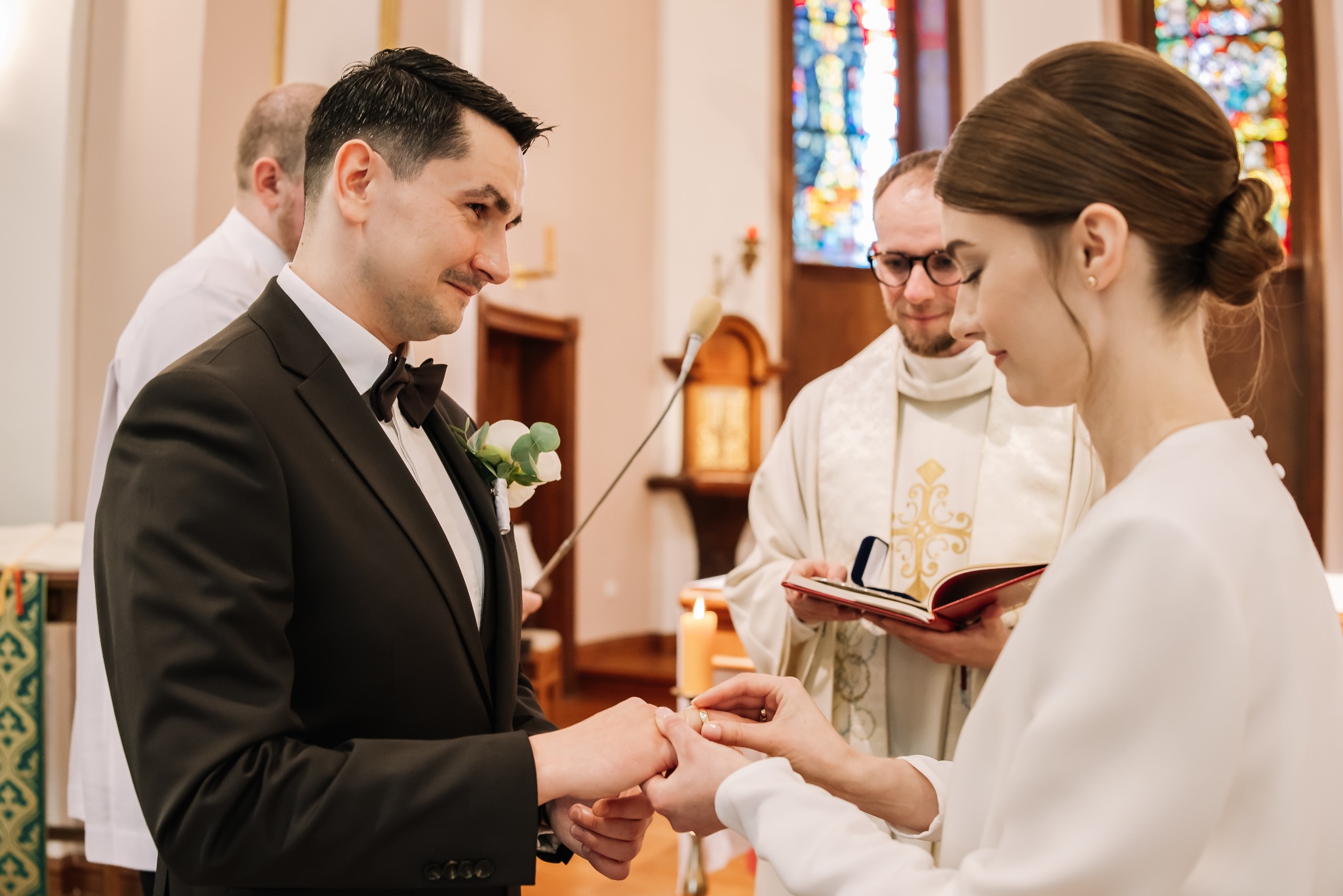 Para młoda wymienia obrączki podczas ceremonii ślubnej w kościele, w tle ksiądz z otwartą księgą. Delikatne światło podkreśla uroczysty charakter chwili.
