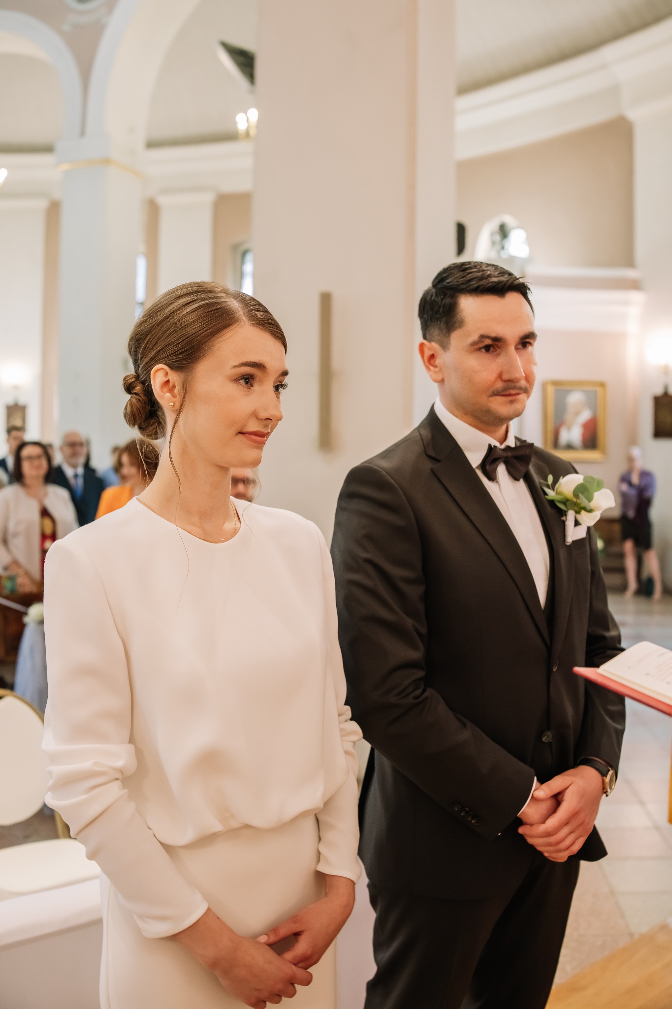 Para młoda w kościele podczas ceremonii ślubnej. Panna młoda w białej sukni, pan młody w czarnym garniturze z muszką. W tle widoczni goście i wnętrze kościoła.