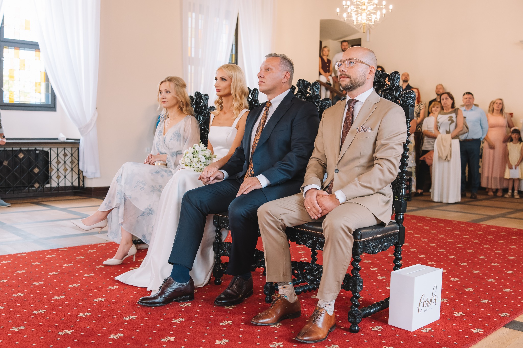 Uroczystość zaślubin w jasnym wnętrzu. Para młoda siedzi obok świadków na zdobionych krzesłach. W tle goście i kryształowy żyrandol, a na podłodze czerwony dywan.