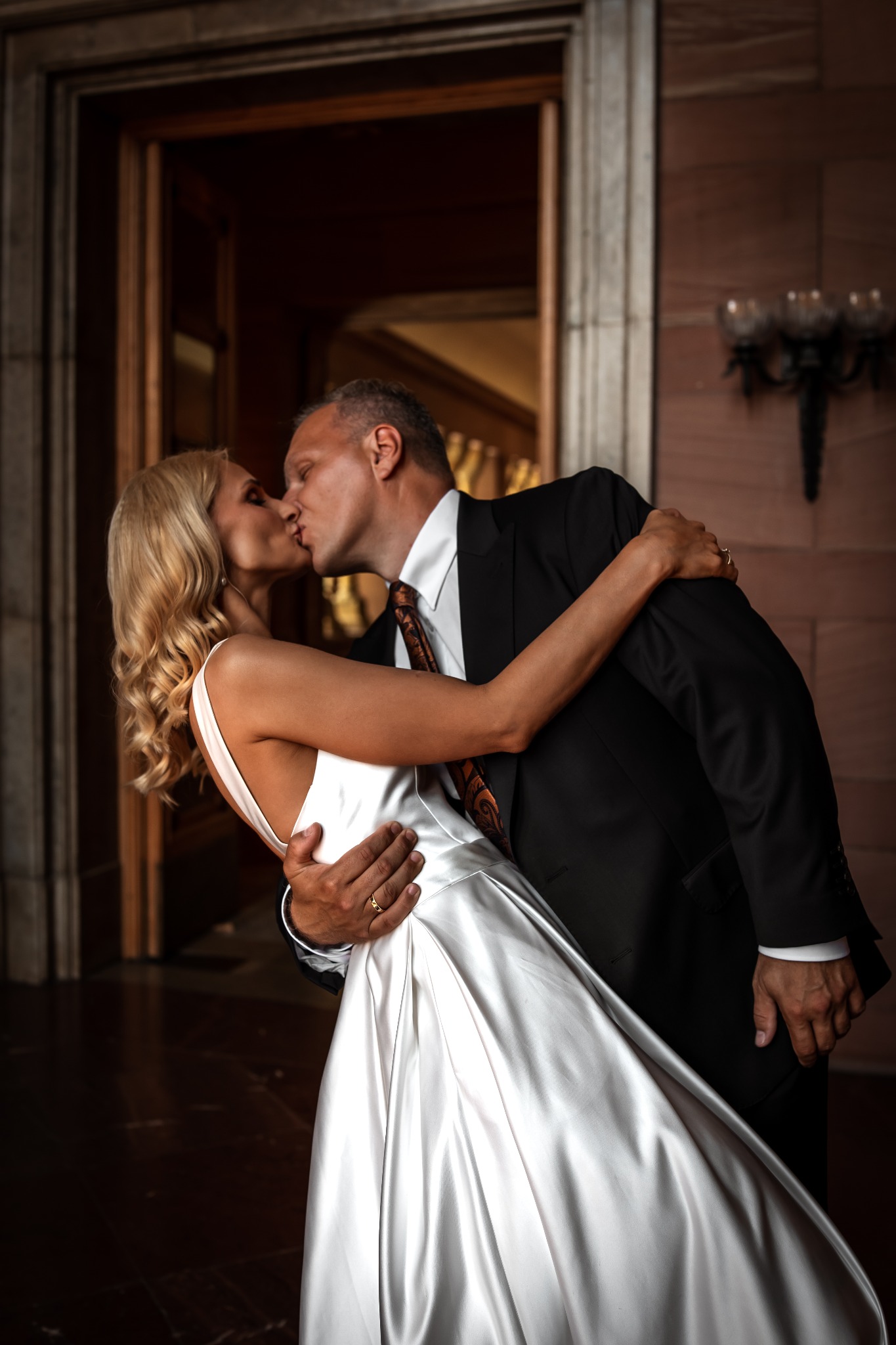 Kadr z ceremonii ślubnej: Panna młoda w białej sukni i pan młody w czarnym garniturze całują się, stojąc na tle klasycznego wnętrza z drewnianymi drzwiami i dekoracyjnymi lampami.