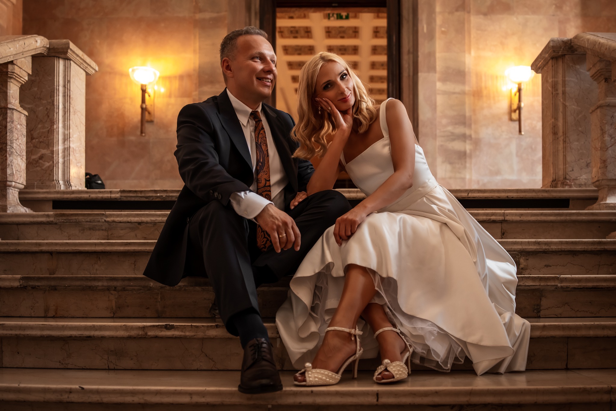 Elegancka para młoda siedzi na marmurowych schodach w ciepłym oświetleniu. Panna młoda w białej sukni i szpilkach, pan młody w garniturze i wzorzystym krawacie.