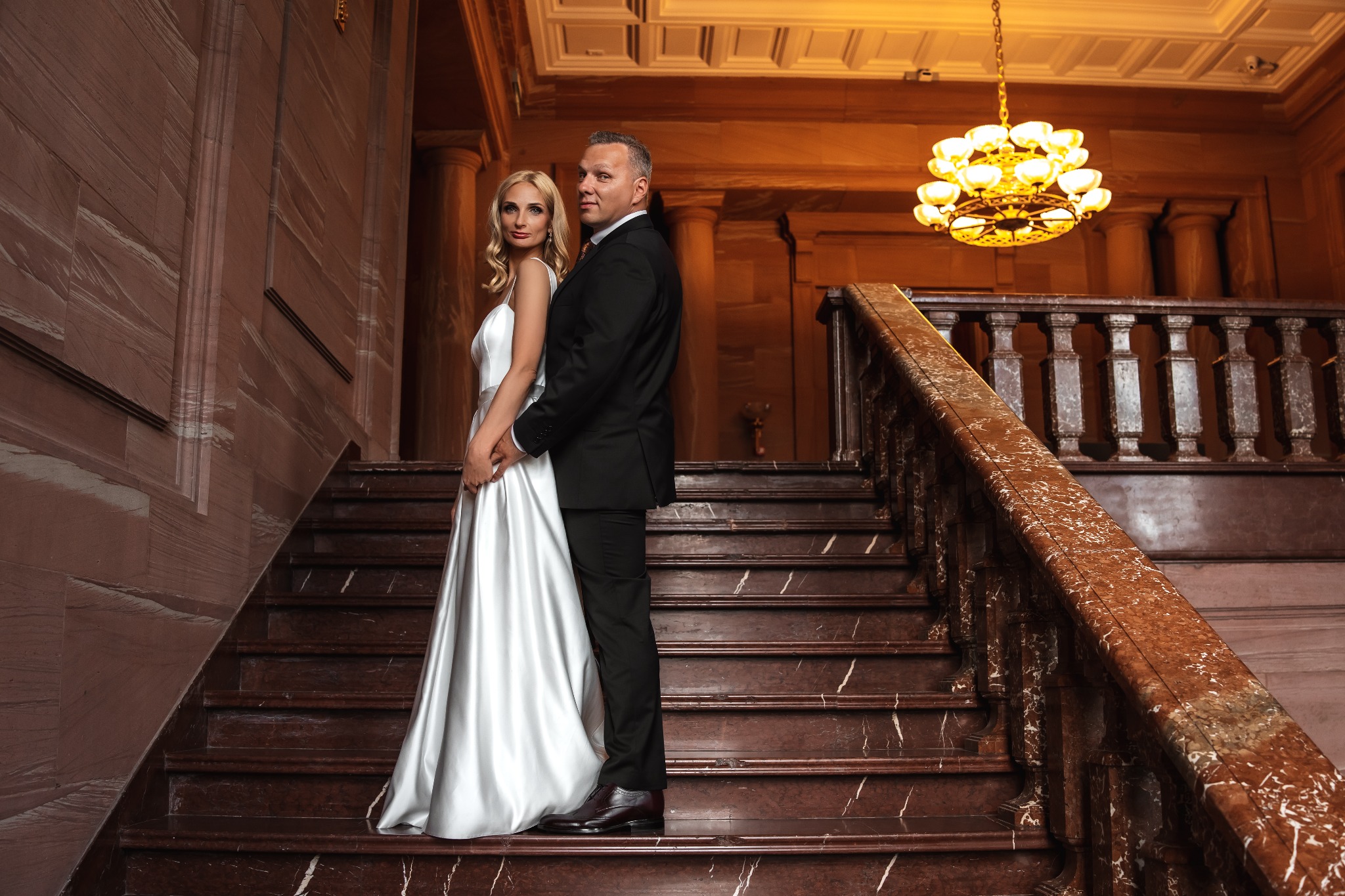 Elegancka para młoda na marmurowych schodach w zabytkowym wnętrzu. Ona w białej sukni, on w czarnym garniturze. W tle kolumny i żyrandol.