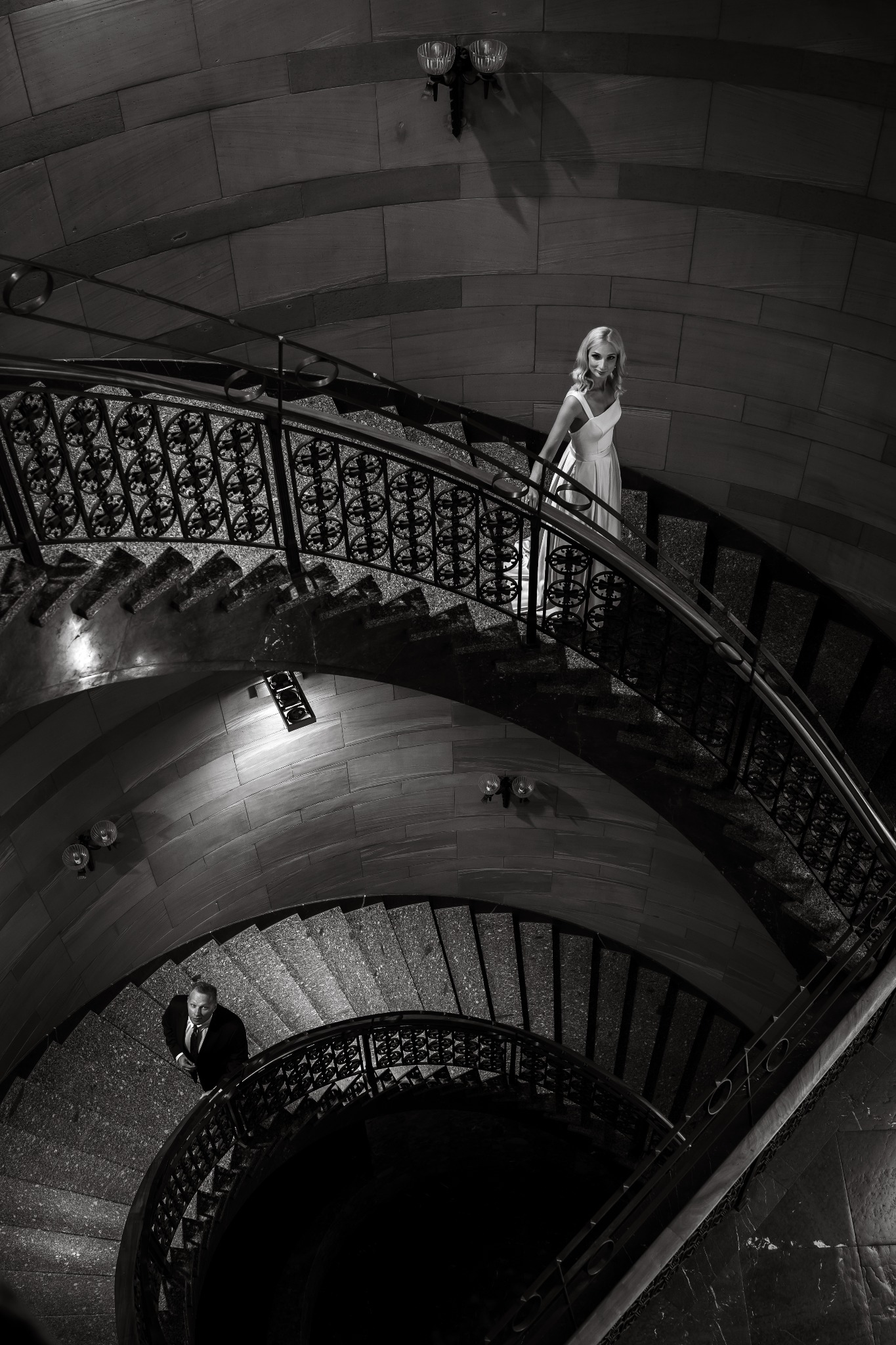 Czarno-białe zdjęcie ślubne z góry: panna młoda w sukni na schodach, pan młody w garniturze spogląda w górę. Dekoracyjne balustrady, oświetlenie podkreśla kamienne wykończenie.