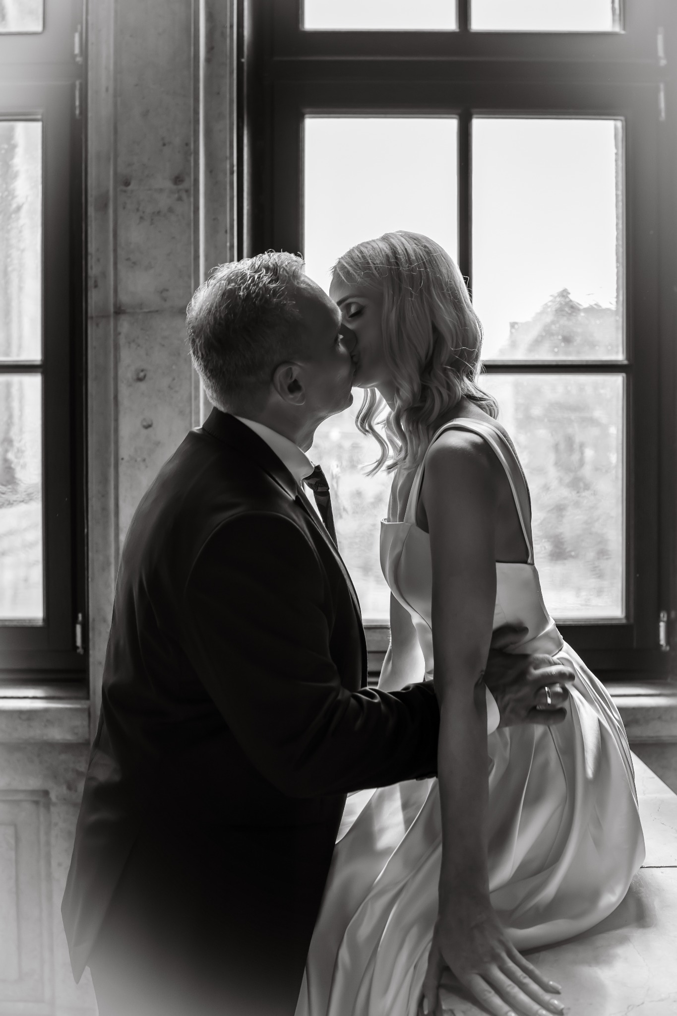 Czarno-białe ujęcie całującej się pary młodej, panna młoda siedzi na parapecie okna, mężczyzna w garniturze stoi obok, delikatne światło okienne.