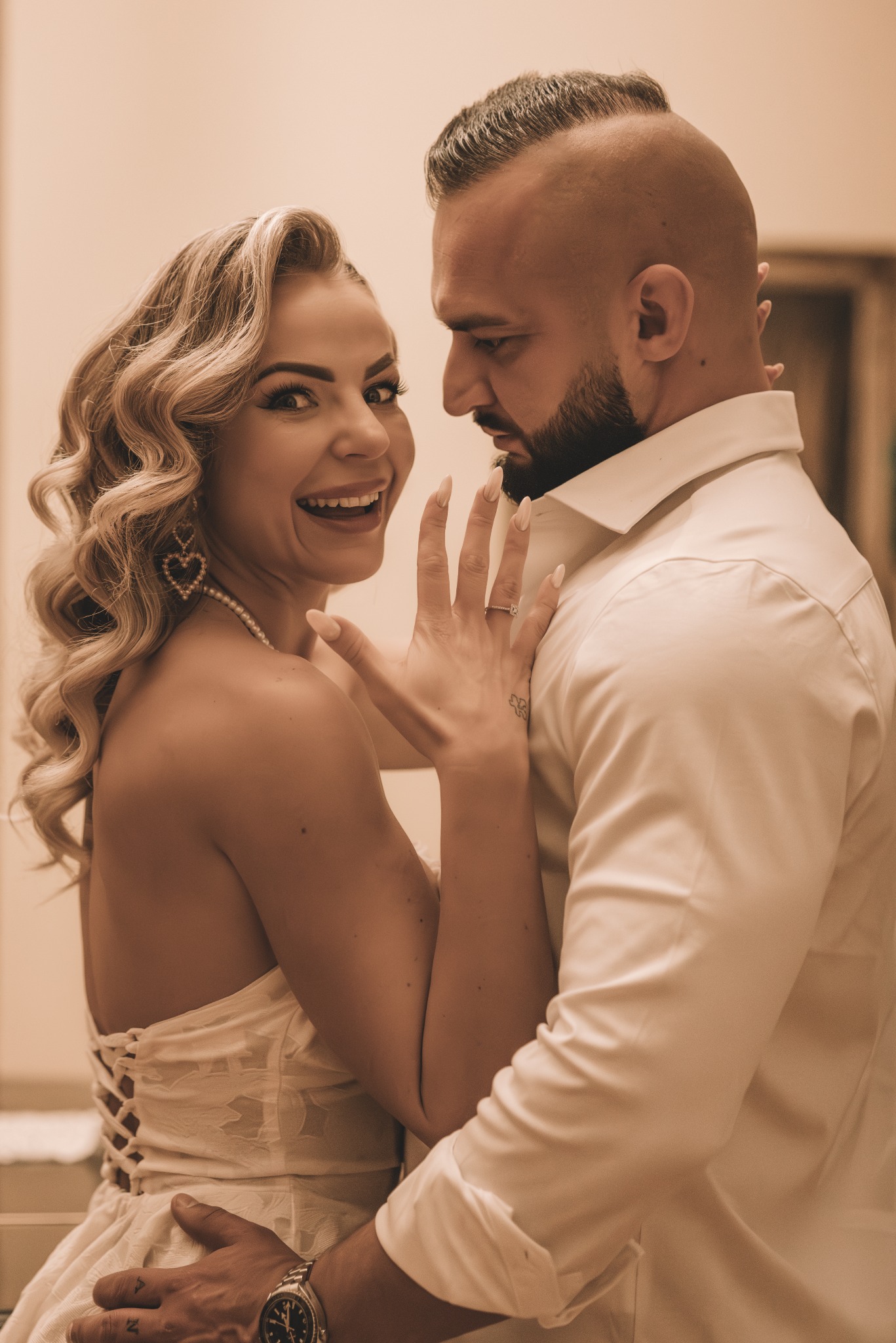 Uśmiechnięta panna młoda prezentuje pierścionek zaręczynowy, obejmowana przez pana młodego w białej koszuli. Romantyczna sesja w ciepłych tonach.