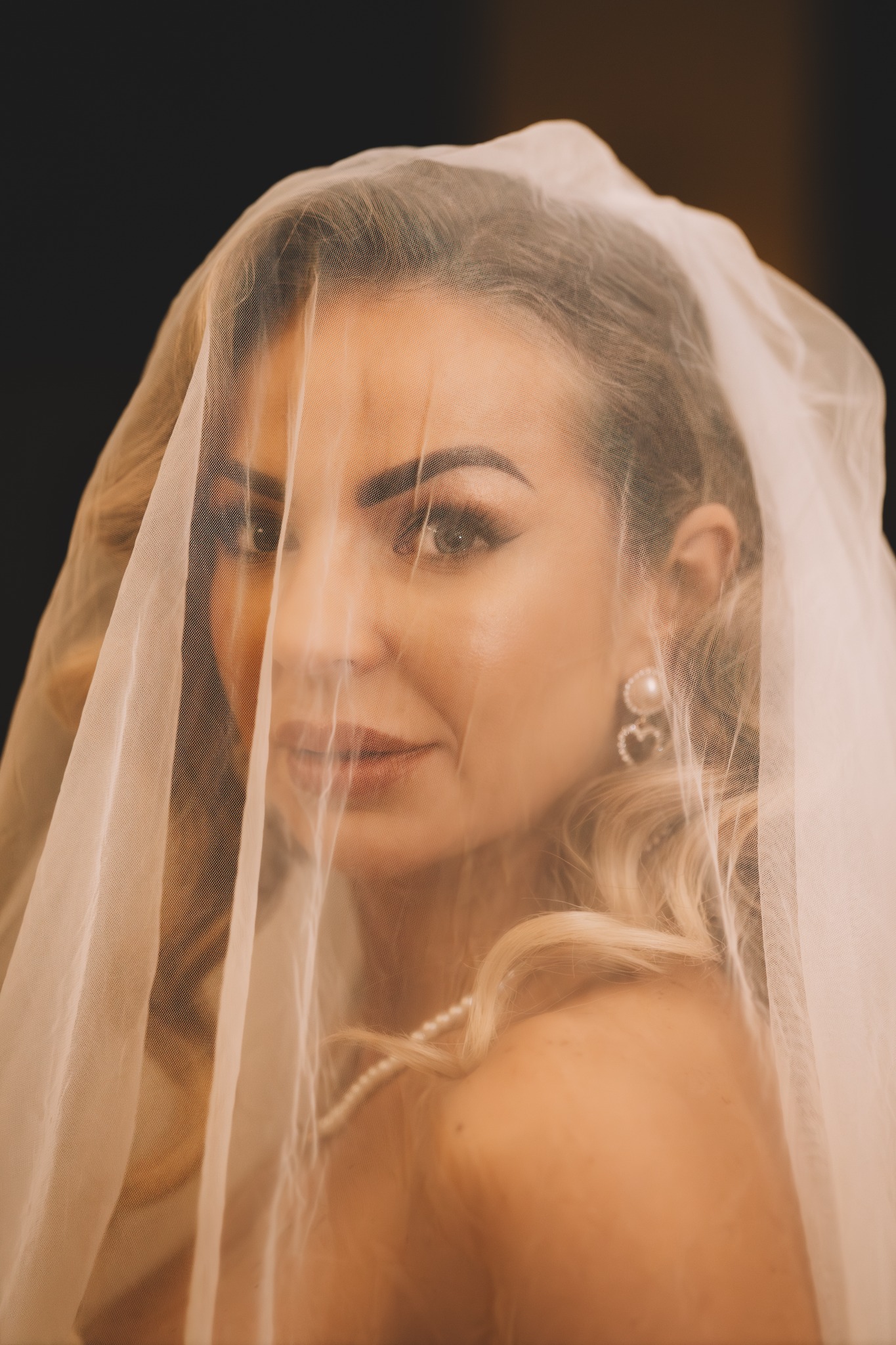 Portret panny młodej z delikatnym makijażem, ukrytej za woalką, z widoczną perłową biżuterią i kolczykami w kształcie serc na ciemnym tle.