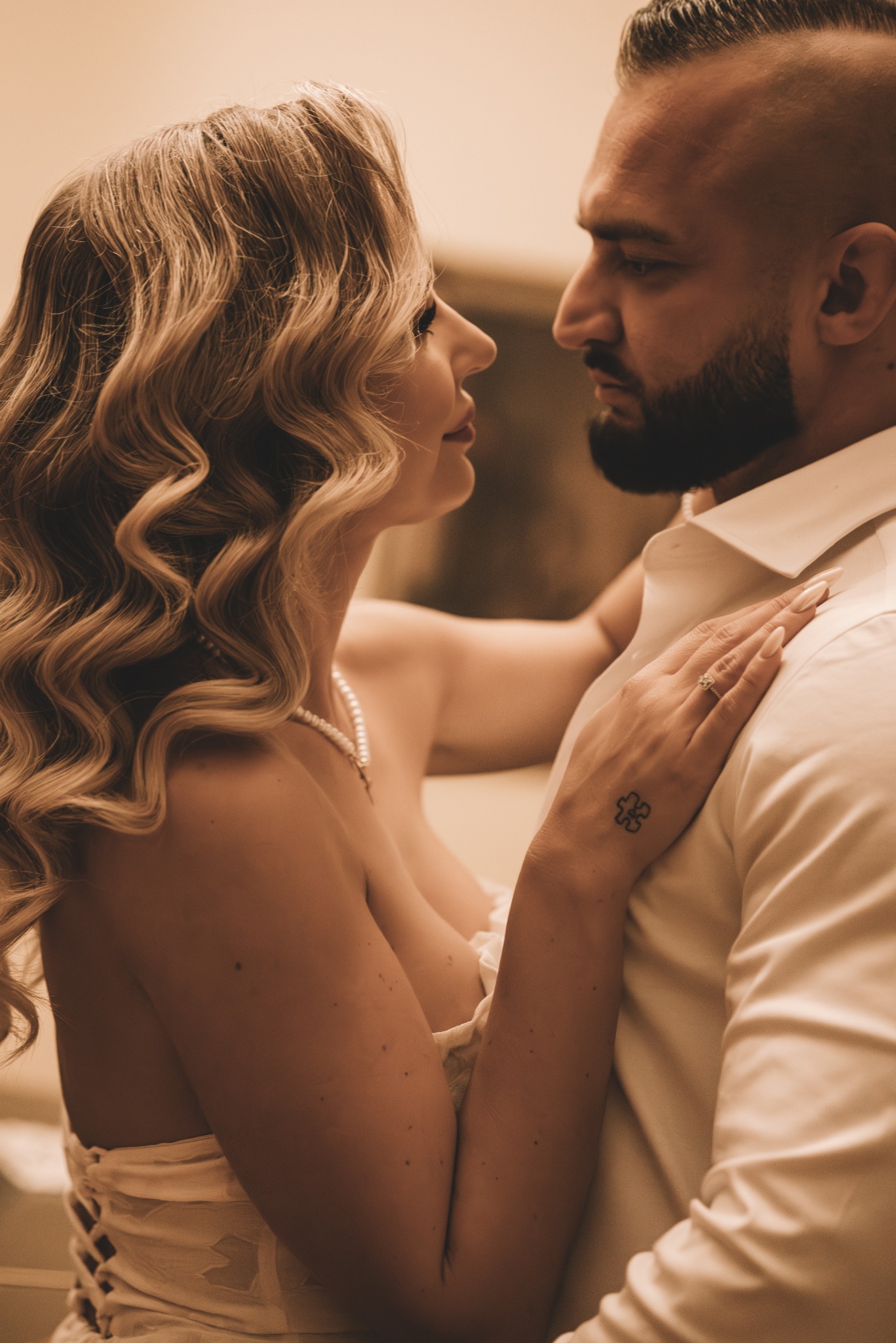 Zmysłowe zbliżenie pary: kobieta z perłami i tatuażem na dłoni opiera się o mężczyznę w białej koszuli, patrząc sobie w oczy. Ciepłe, stonowane barwy.