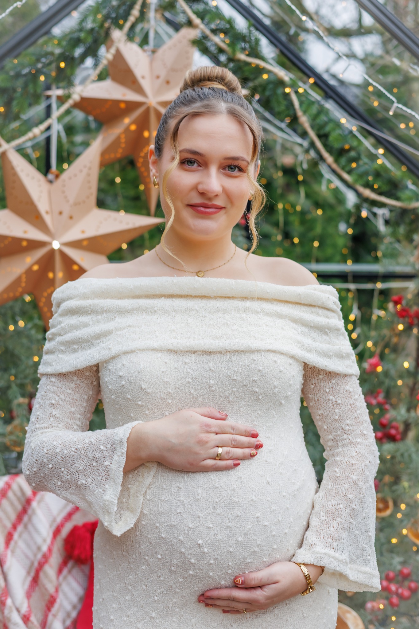Portret kobiety w ciąży w białej sukni, dłonie na brzuchu, w tle dekoracje świąteczne i gwiazdy. Uśmiechnięta przyszła mama w zimowej scenerii.