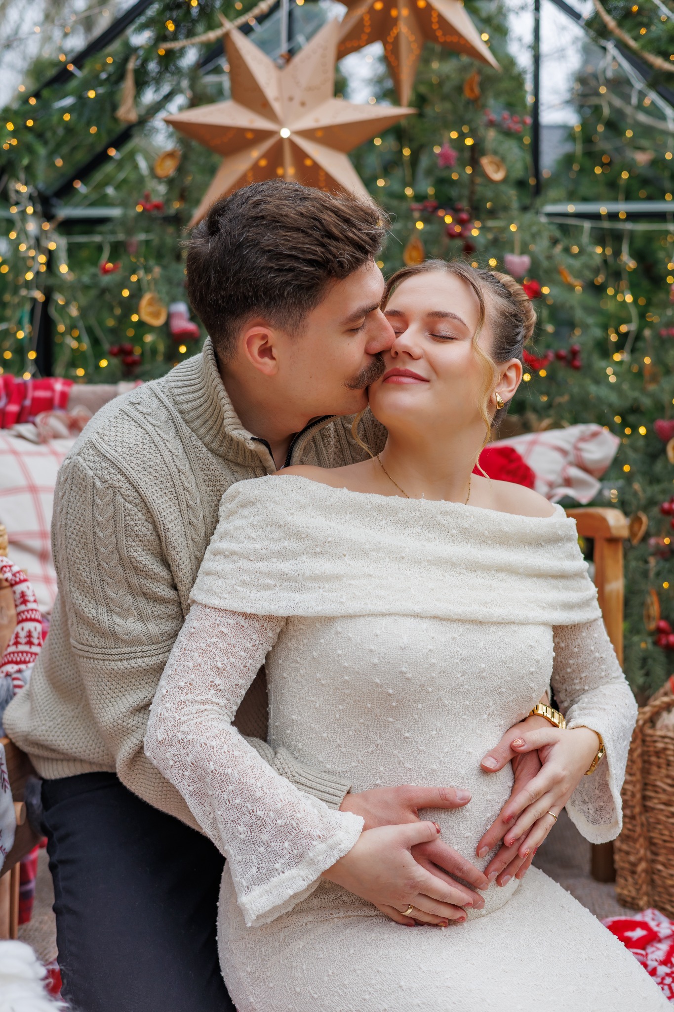 Czuły portret przyszłych rodziców w świątecznej scenerii: mężczyzna całuje kobietę w ciążowej sukience, dłonie ułożone na brzuchu w kształt serca, ciepłe oświetlenie.
