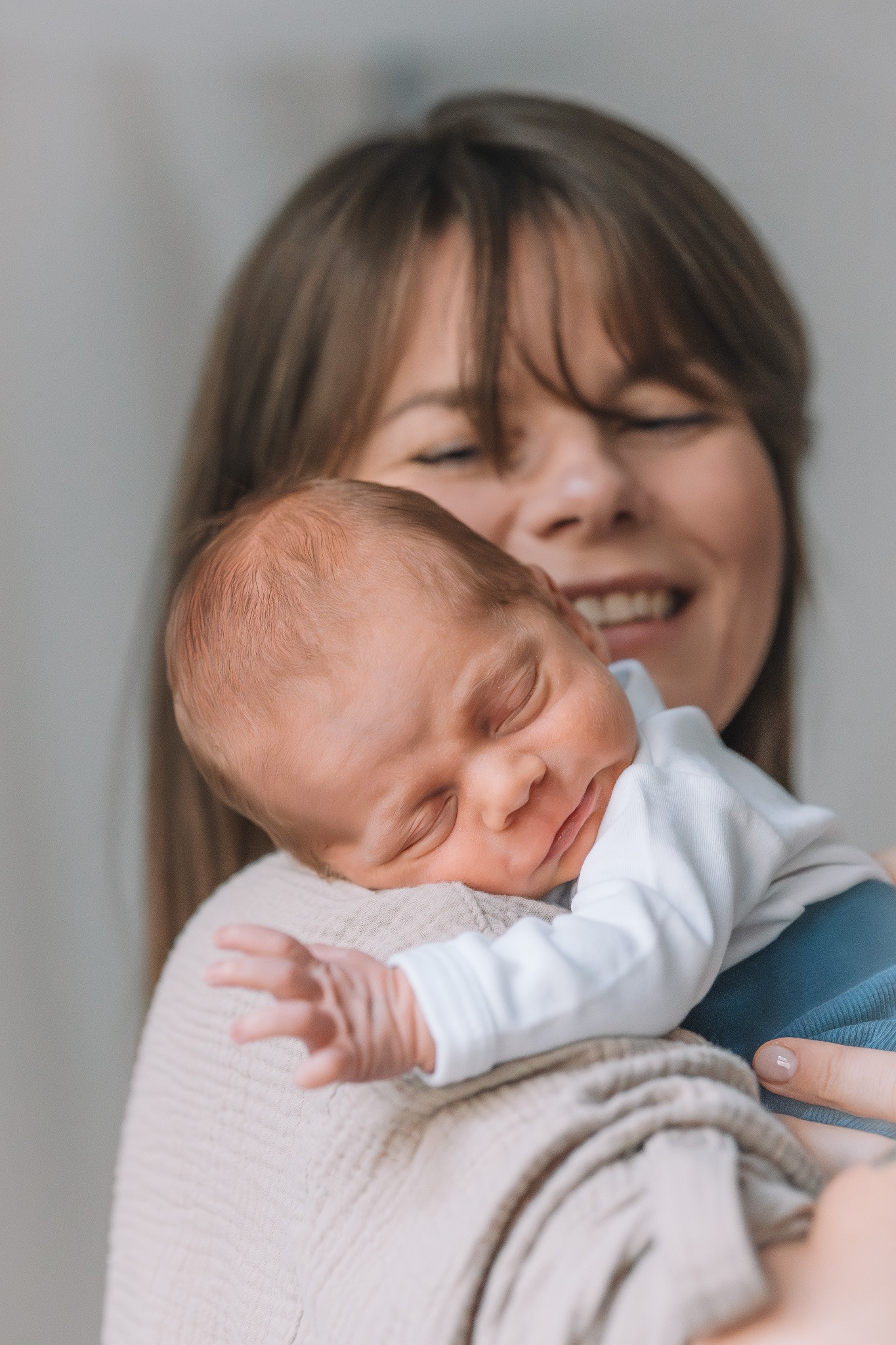 Mama trzyma na ramieniu śpiącego noworodka w białym body. Kobieta uśmiecha się, patrząc na dziecko. Delikatne, naturalne światło podkreśla bliskość i czułość.