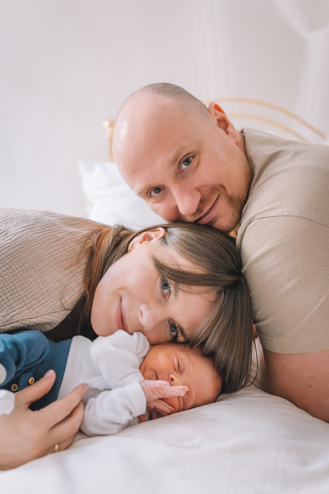 Rodzinne ujęcie: mama, tata i noworodek leżący na białej pościeli, blisko siebie, z delikatnym uśmiechem, w jasnym, naturalnym świetle.