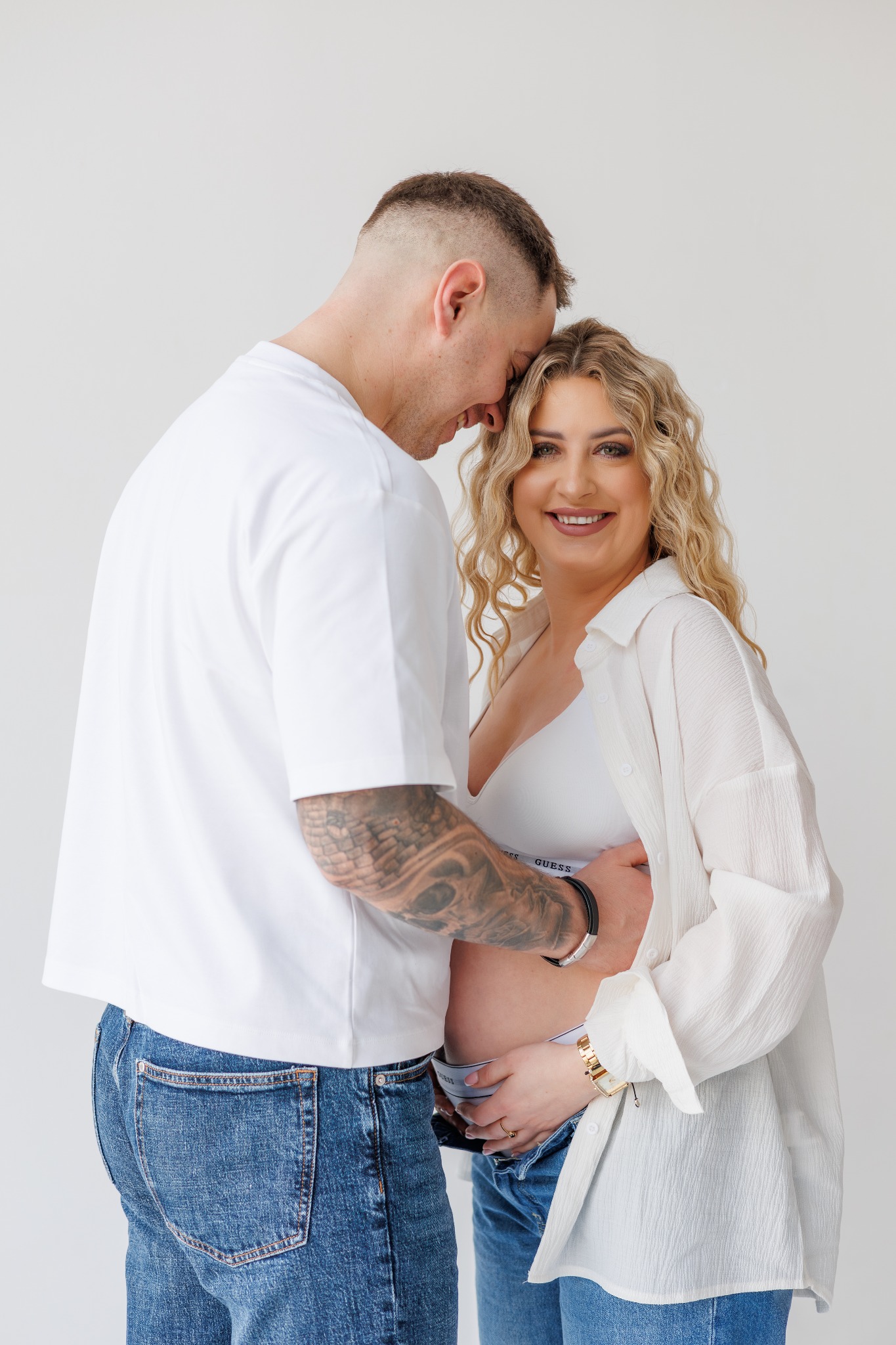 Uśmiechnięta, ciężarna blondynka w białej koszuli, obejmowana przez mężczyznę z tatuażami na ramieniu, oboje w jeansach, na jasnym tle.