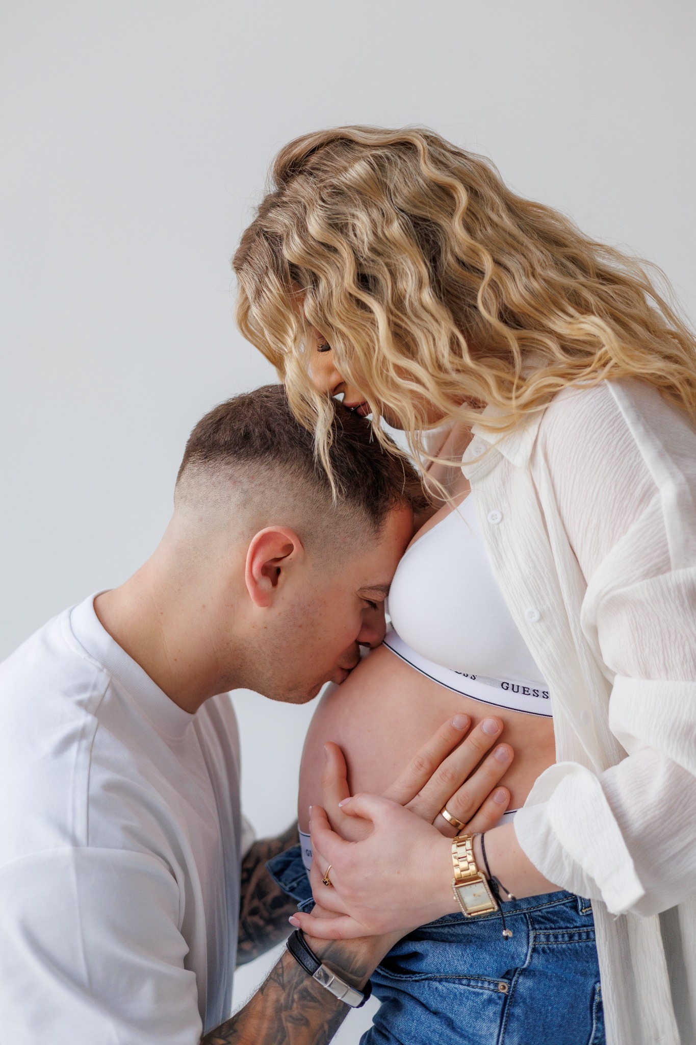 Mężczyzna całuje ciążowy brzuch kobiety w bieliźnie Guess i rozpiętej koszuli. Jasne tło, delikatne światło, bliski kadr, widoczne tatuaże na ręce mężczyzny.