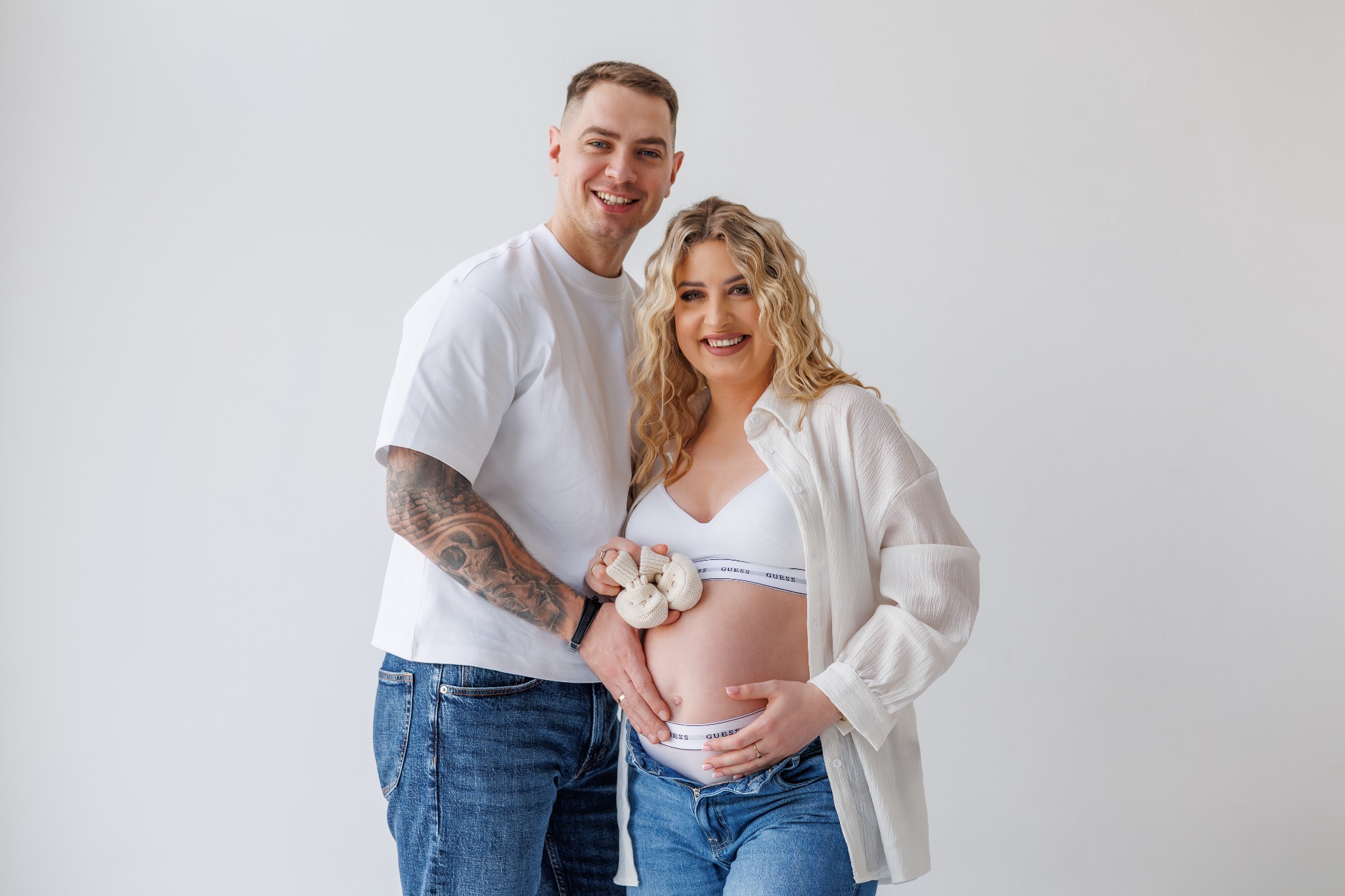 Uśmiechnięta para w jasnych ubraniach pozuje na sesji ciążowej. Kobieta trzyma małe, dziergane buciki na brzuchu. Mężczyzna ma tatuaż na ramieniu. Białe tło.