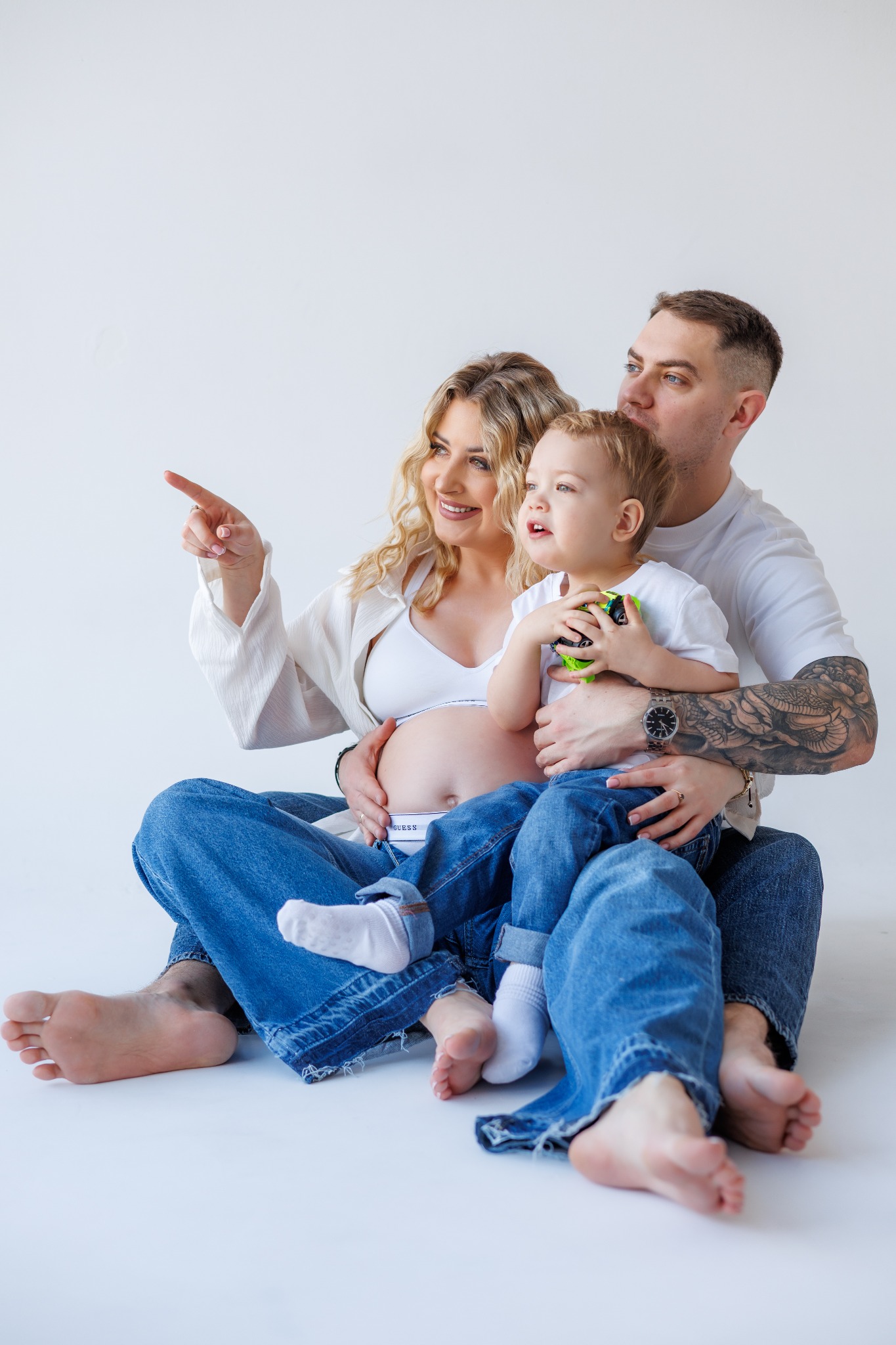 Uśmiechnięta, przyszła mama z blond lokami, tata z tatuażem i synek siedzą na białym tle. Kobieta wskazuje palcem w dal. Sesja w jeansach i bieli.