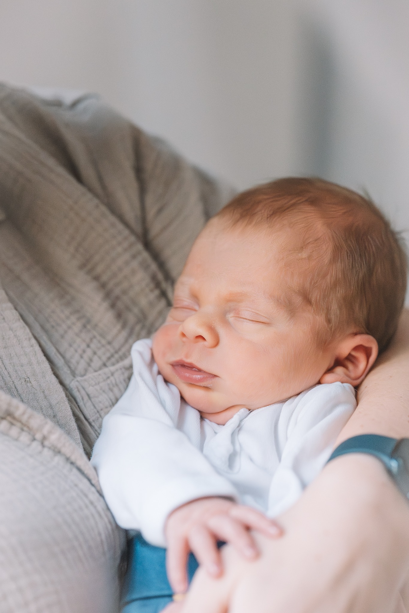 Śpiący noworodek w białym ubranku, trzymany w ramionach osoby w beżowej koszuli. Delikatne światło podkreśla rysy twarzy dziecka.