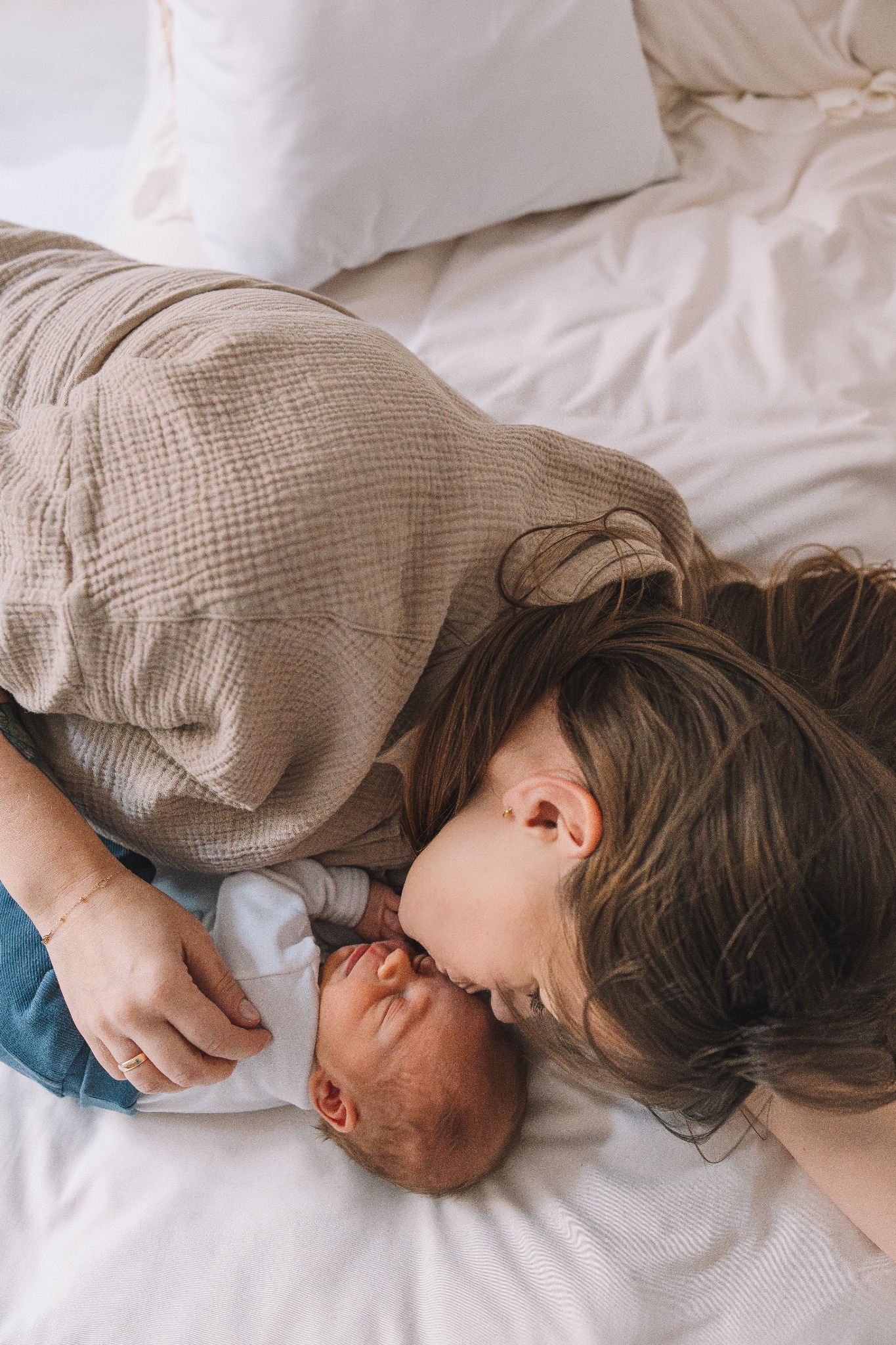 Matka w beżowej bluzce całuje śpiącego noworodka w białym ubranku, leżąc na białej pościeli. Delikatne światło podkreśla intymność i bliskość relacji.