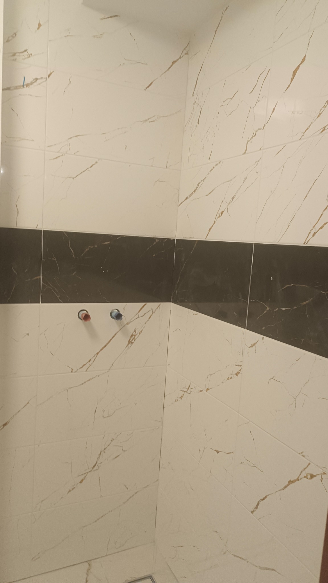 Wykończona ściana w łazience z białych płytek imitujących marmur z poziomym pasem ciemnych płytek, widoczne wystające rury instalacji wodnej z zaślepkami w kolorze czerwonym i niebieskim.