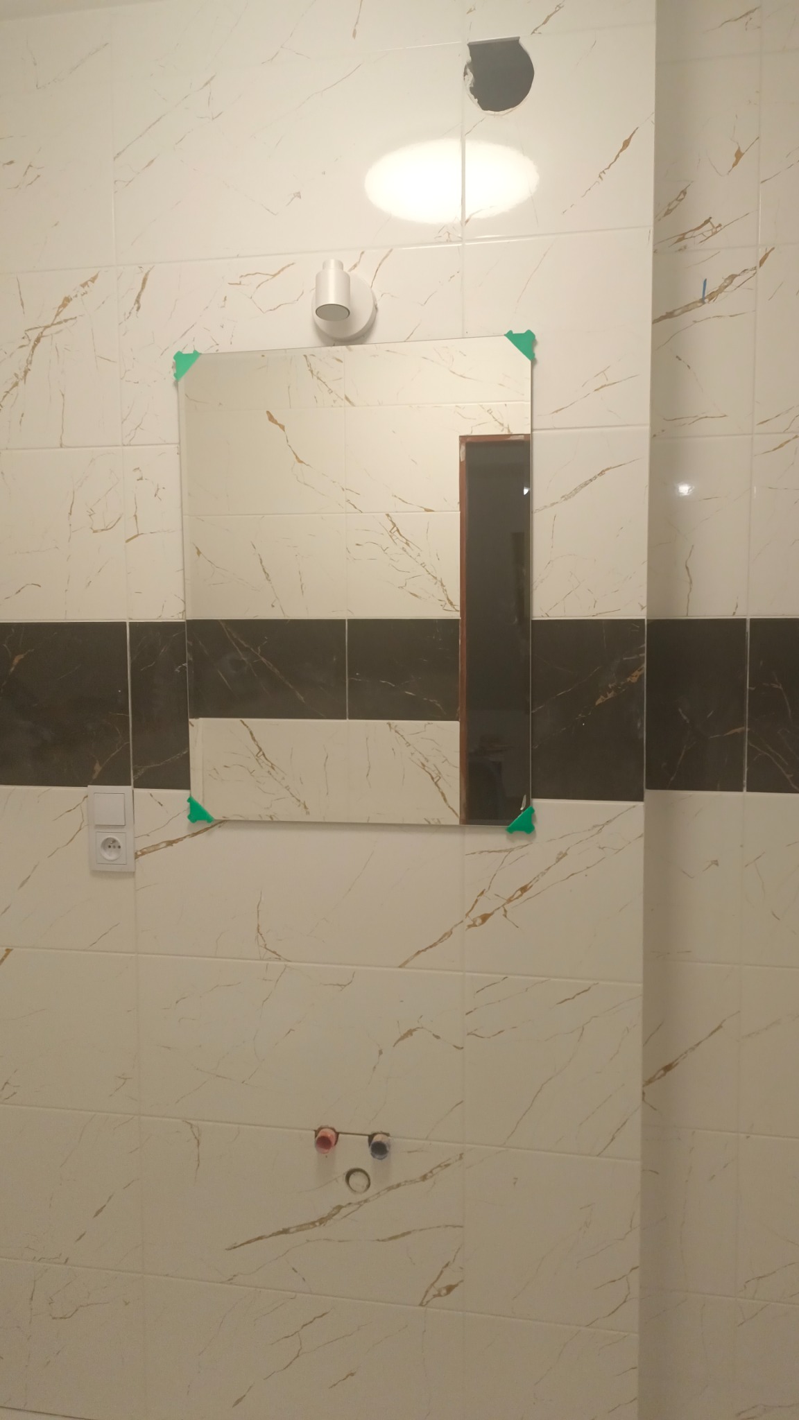 Ściana łazienki w trakcie wykańczania z zamontowanym lustrem, zabezpieczonym narożnikami, widoczne przyłącza wodne i otwór na oświetlenie w płytkach imitujących marmur.