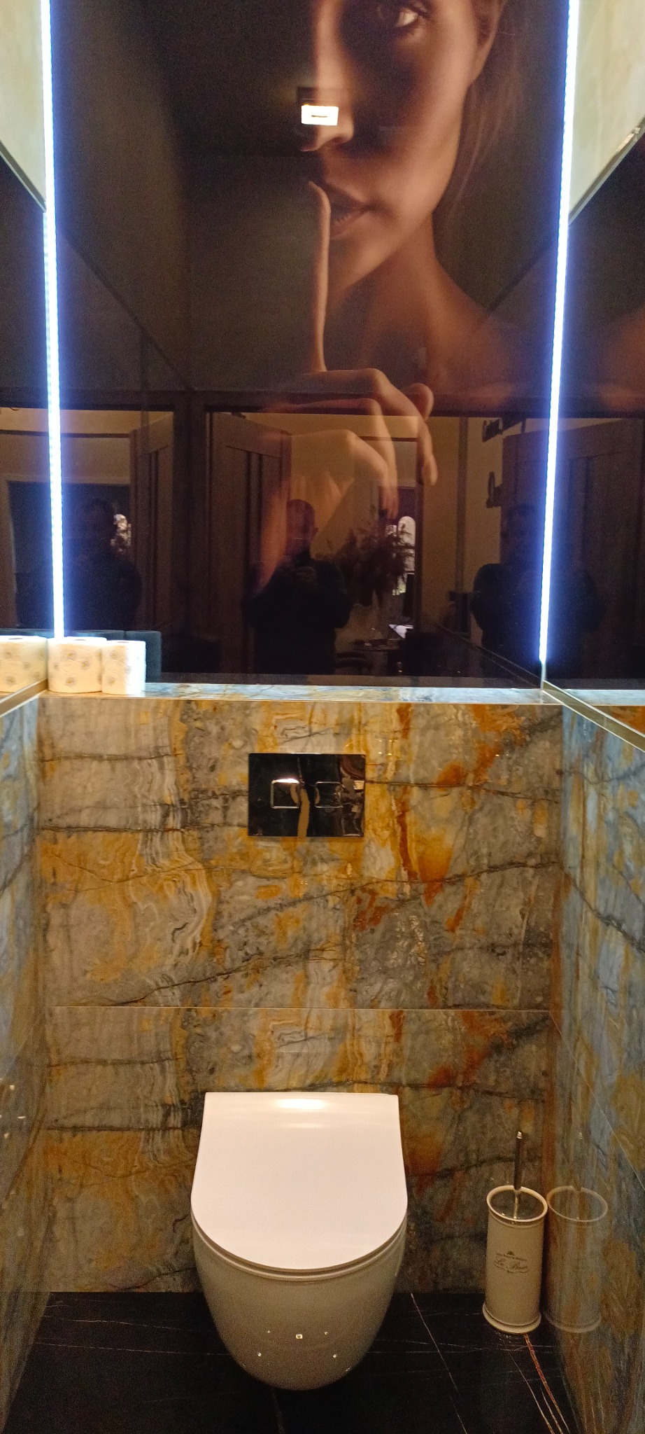 Nowoczesna toaleta wisząca w łazience wykończonej płytkami imitującymi marmur z pomarańczowymi żyłami, na suficie podwieszanym lustro z podświetleniem LED, odbijające fragment twarzy kobiety...