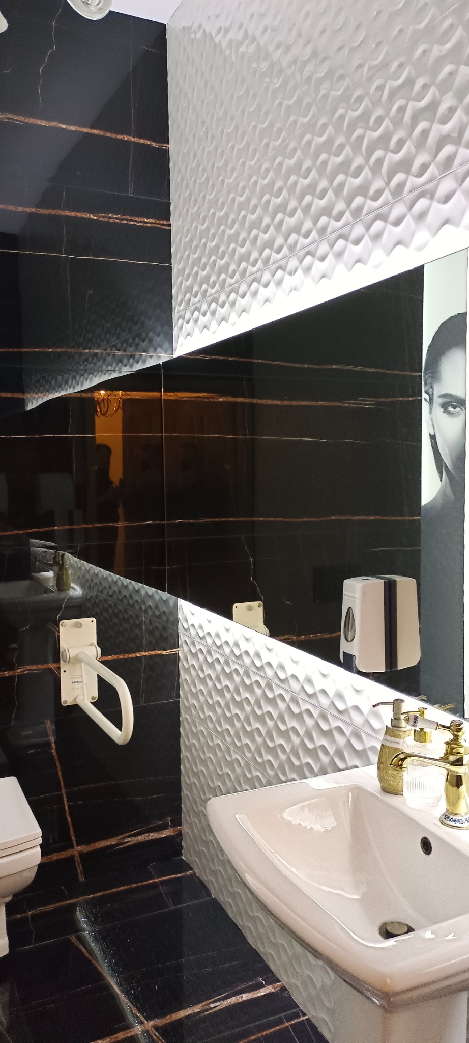 Wykończona łazienka z czarnymi płytkami imitującymi marmur z złotymi żyłami, kontrastującymi z białą, teksturowaną ścianą, widoczna umywalka, toaleta z uchwytem dla niepełnosprawnych i lustro...