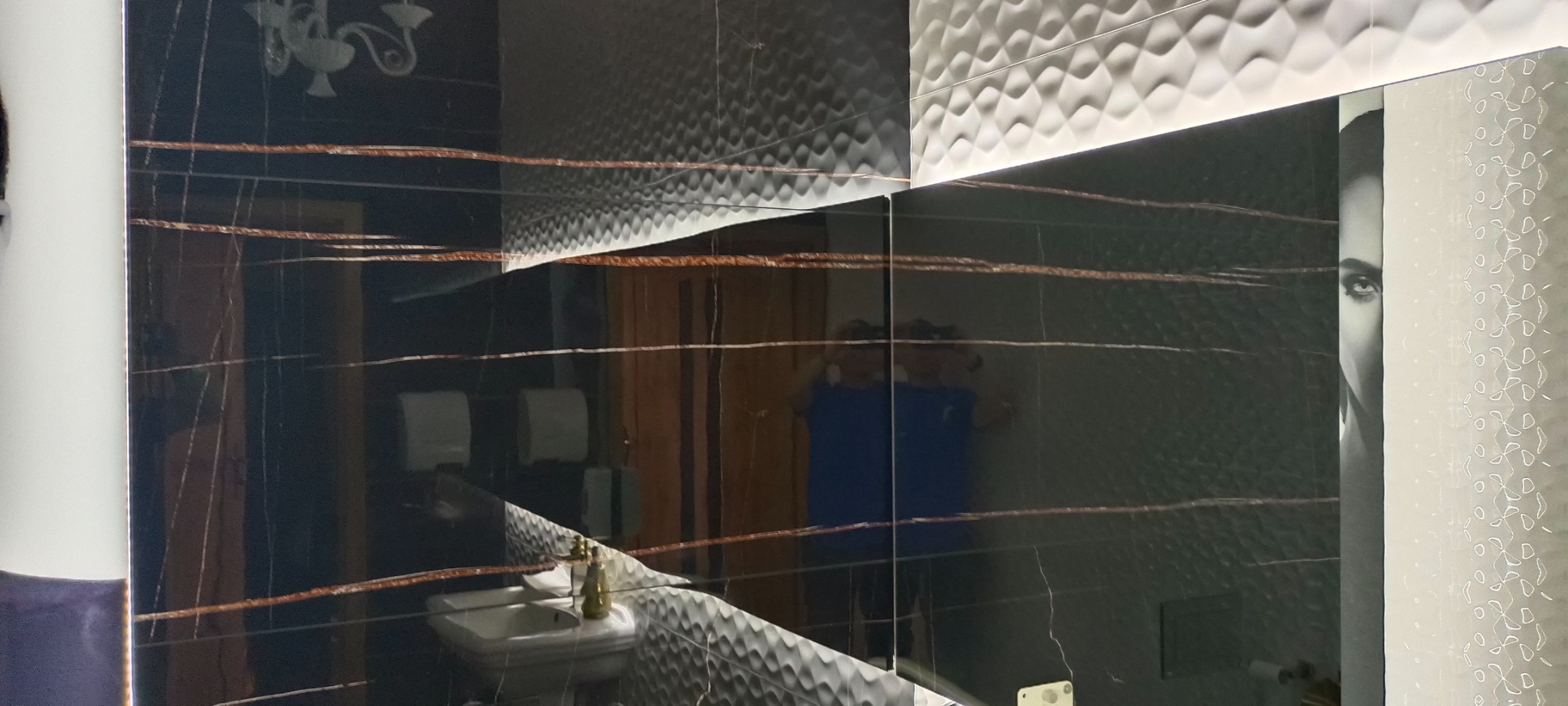 Wykończona łazienka z czarnymi płytkami imitującymi marmur z brązowymi żyłami, dekoracyjnymi białymi płytkami 3D i lustrem z grafiką twarzy kobiety.