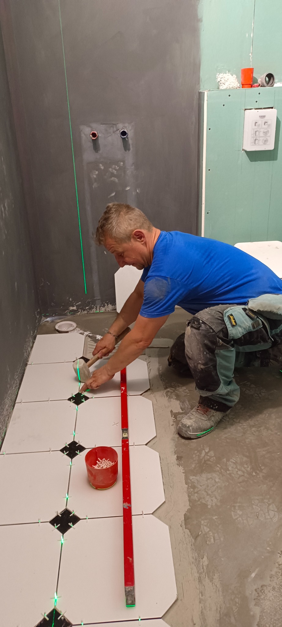 Fachowiec w trakcie układania białych płytek podłogowych z czarnymi akcentami w łazience, używając poziomicy laserowej i młotka gumowego, widoczne krzyżyki dystansowe i czerwone wiaderko z klinami.