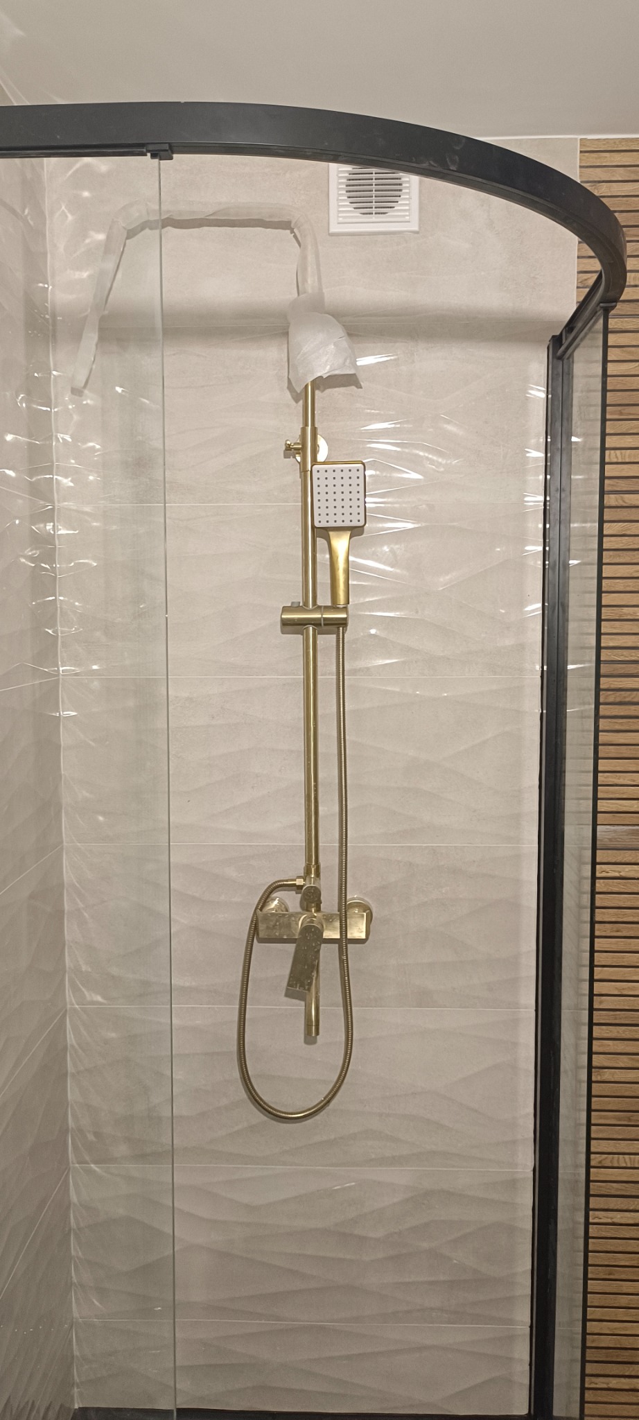 Nowa, złota bateria prysznicowa z deszczownicą, zabezpieczona folią ochronną, zamontowana na ścianie z trójwymiarowymi, jasnymi płytkami w kabinie prysznicowej z czarnymi profilami i drewnianymi...