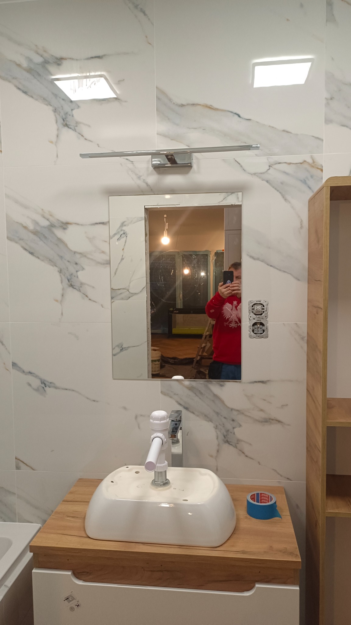 Łazienka w trakcie remontu: białe płytki imitujące marmur, zamontowane lustro z oświetleniem, umywalka bez baterii na drewnianym blacie, widoczne puszki elektryczne bez osprzętu, w lustrze odbicie...