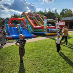 Kolorowy, dmuchany plac zabaw z motywem bajki 'Psi Patrol', zjeżdżalnie oraz dzieci bawiące się na trawie w słoneczny dzień.