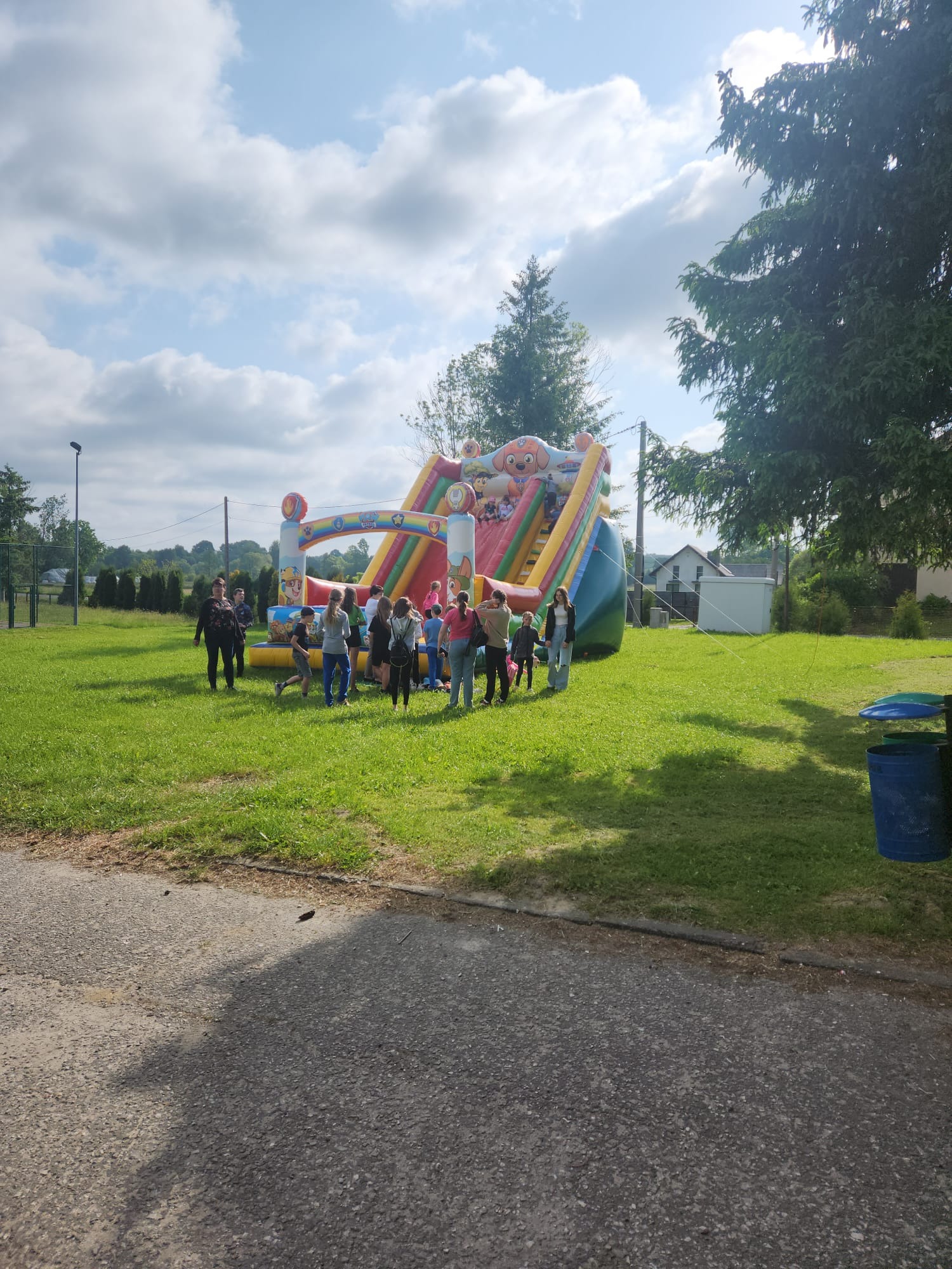 Dmuchany zamek z motywem bajki Psi Patrol, ustawiony na trawie, otoczony grupą dzieci i dorosłych w słoneczny dzień.