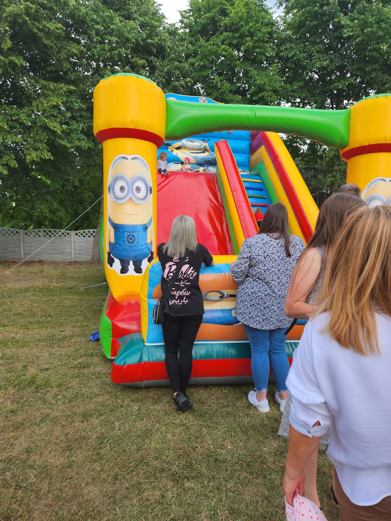 Kolorowy dmuchany zamek ze zjeżdżalnią na trawie, z motywem postaci z bajki, obserwowany przez grupę osób dorosłych z tyłu.